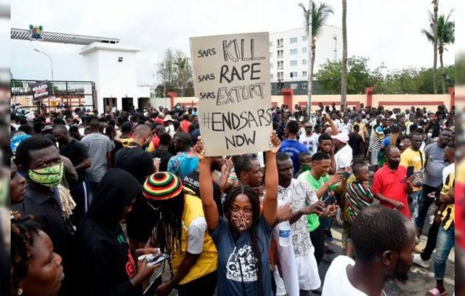 The image depicts a large crowd of people gathered in a protest setting. One individual is holding a sign that reads "KILL RAPE EXTORT END SARS NOW." The atmosphere appears to be serious and unified, with many participants wearing masks. The background includes some trees and buildings, indicating an outdoor urban environment. The protest is likely focused on social issues related to law enforcement.