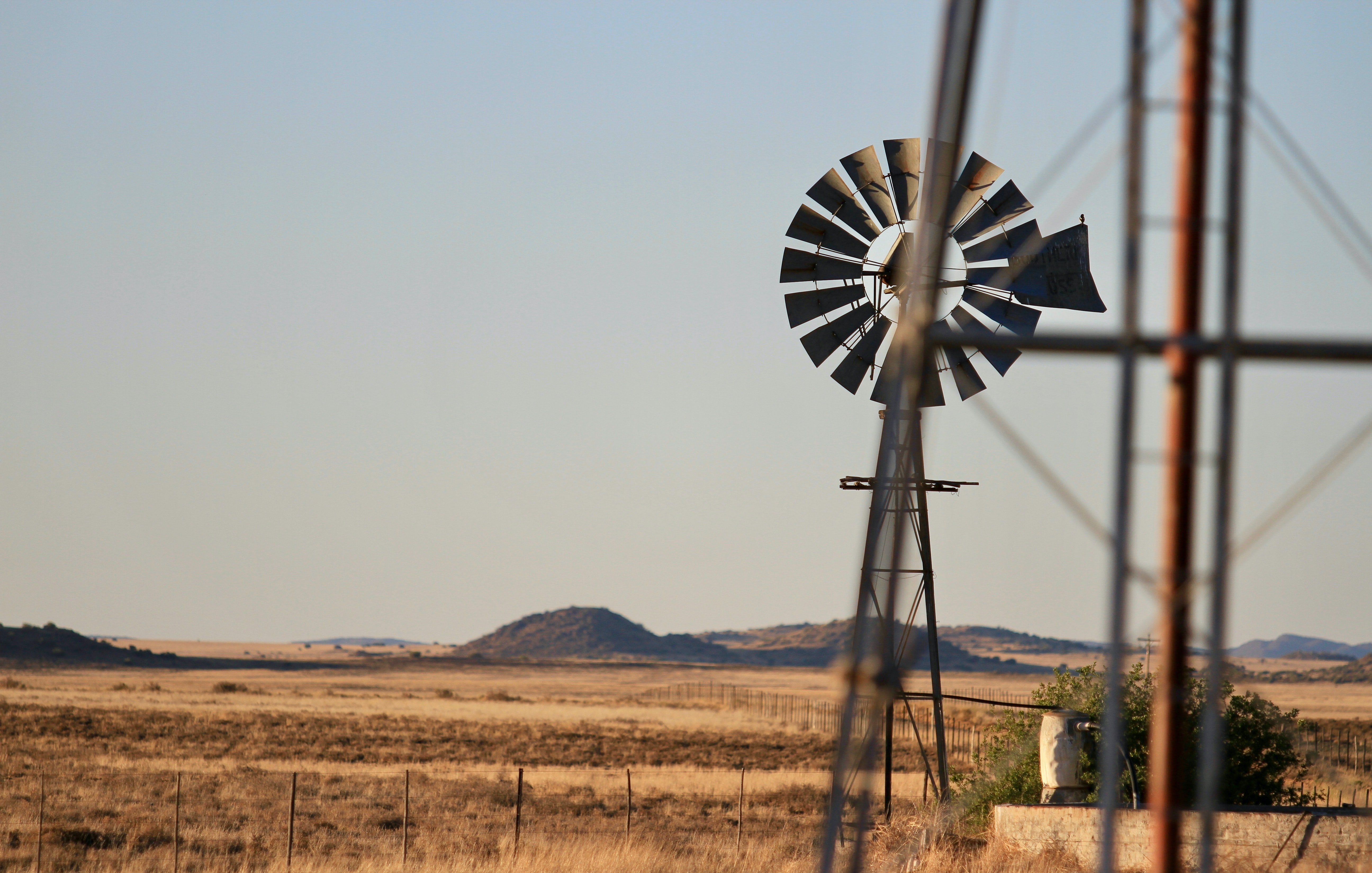 L'image montre un paysage rural étendu sous un ciel dégagé. À l'arrière-plan, des collines douces se dessinent sur l'horizon, tandis que le sol est recouvert d'une herbe sèche, témoignant d'un environnement aride. Au premier plan, une éolienne en métal tourne doucement, projetant son ombre sur le sol. Les pales de l'éolienne sont bien visibles, capturant quelques rayons de soleil. À proximité, un pilier métallique se dresse légèrement flou, ajoutant une touche de profondeur à la scène. L'ensemble évoque une atmosphère calme et tranquille, comme un moment suspendu dans la nature.