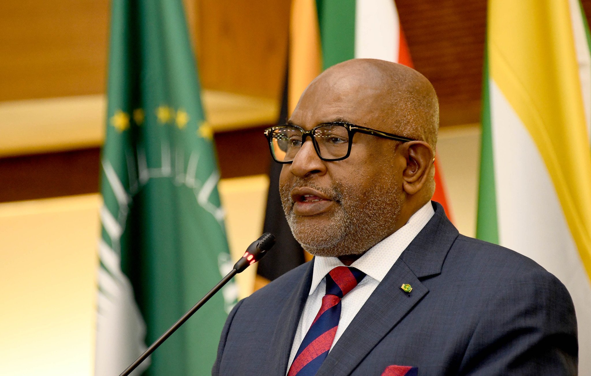 L'image représente un homme en train de prendre la parole lors d'un événement officiel. Il porte un costume sombre avec une chemise blanche et une cravate rayée, dans des tons de rouge et bleu. Ses lunettes sur le nez lui donnent une allure distinguée. Il se tient près d'un microphone, semblant s'adresser à un public. En arrière-plan, on peut apercevoir des drapeaux représentant différents pays, ce qui suggère que l'événement a une portée internationale. L'atmosphère semble solennelle et officielle, marquée par un éclairage chaleureux.