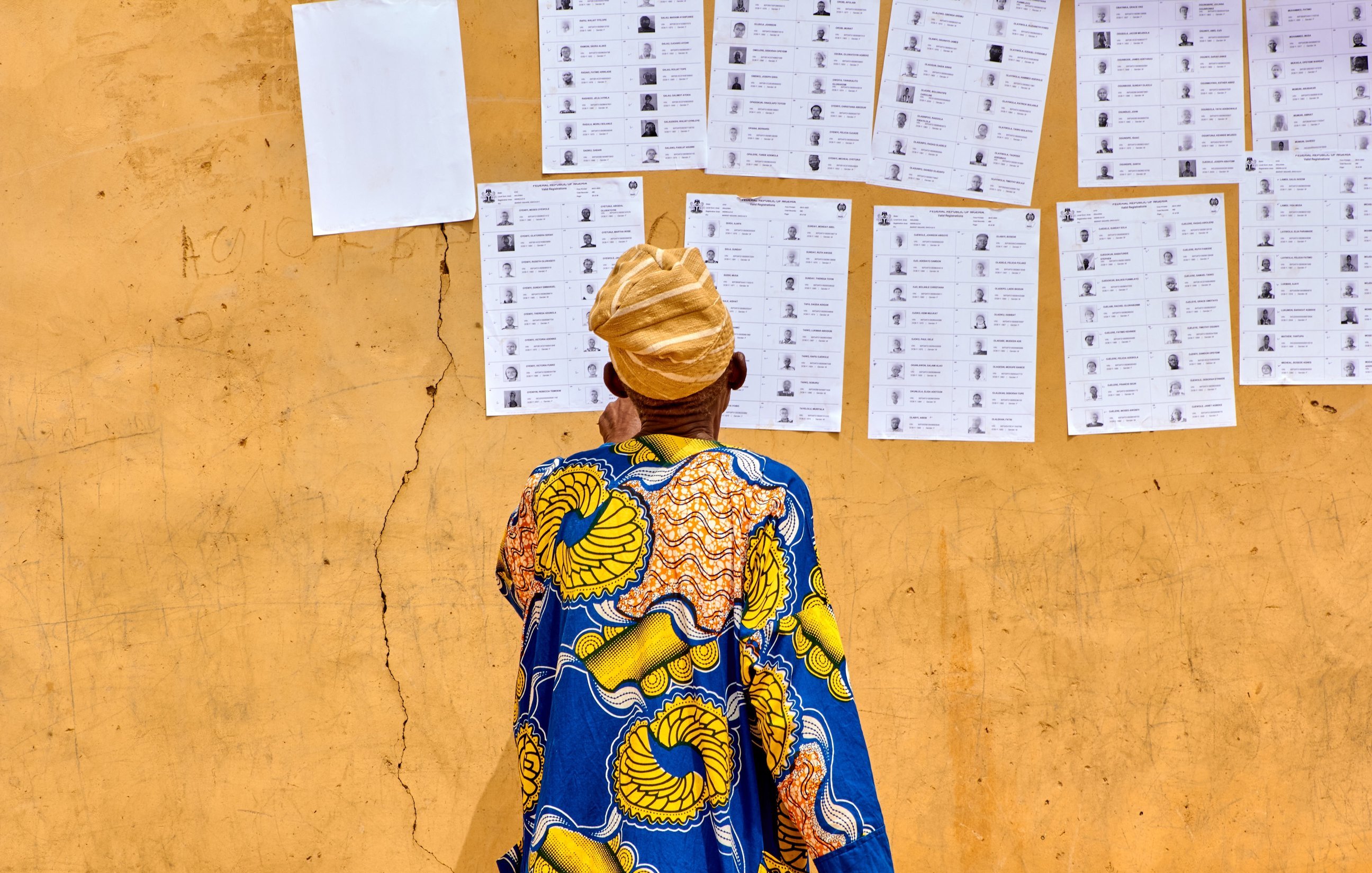 L'image montre un homme de dos, vêtu d'une robe colorée avec des motifs traditionnels, principalement en bleu et jaune. Il se tient devant un mur de couleur ocre, où sont affichés plusieurs documents. Ces documents ressemblent à des bulletins de vote ou des listes, alignés verticalement, avec des photographies et des noms inscrits. Le sol est légèrement fissuré, ajoutant une texture au fond de l'image. L'homme porte un turban beige, et son attention semble focalisée sur les informations affichées sur le mur.