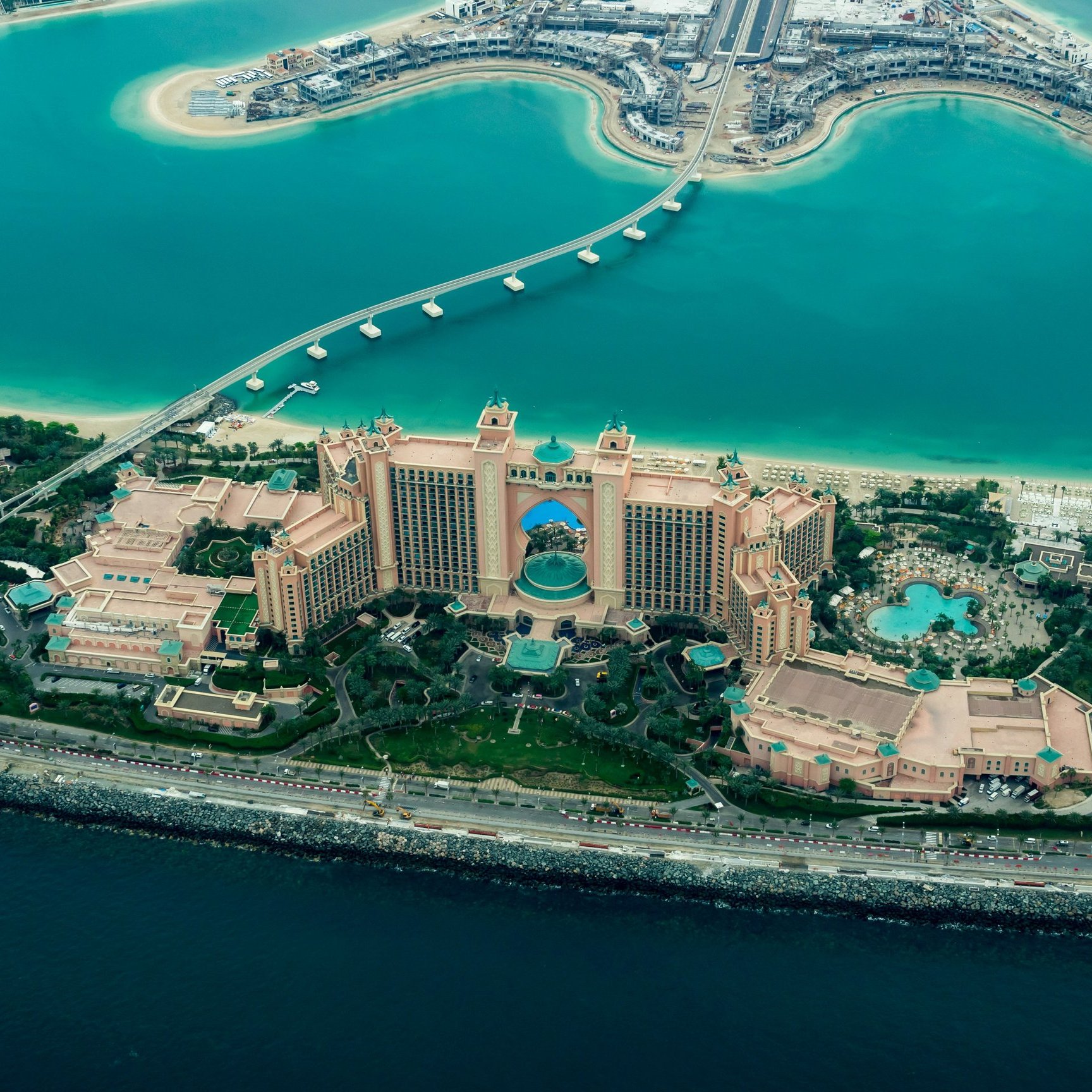 Cette image offre une vue aérienne d'un majestueux complexe hôtelier, entouré par des eaux turquoise. Le bâtiment principal, qui semble imposant, est décoré de nuances de beige et d'or, avec une architecture inspirée des contes arabes. Des jardins luxuriants entourent l'hôtel, agrémentés de piscines scintillantes et de palmiers. À proximité, on peut voir un chemin qui s'étend au-dessus de l'eau, reliant le terre-plein à d'autres structures. L'ensemble dégage une atmosphère luxueuse et tropicale, évoquant des vacances chaleureuses.
