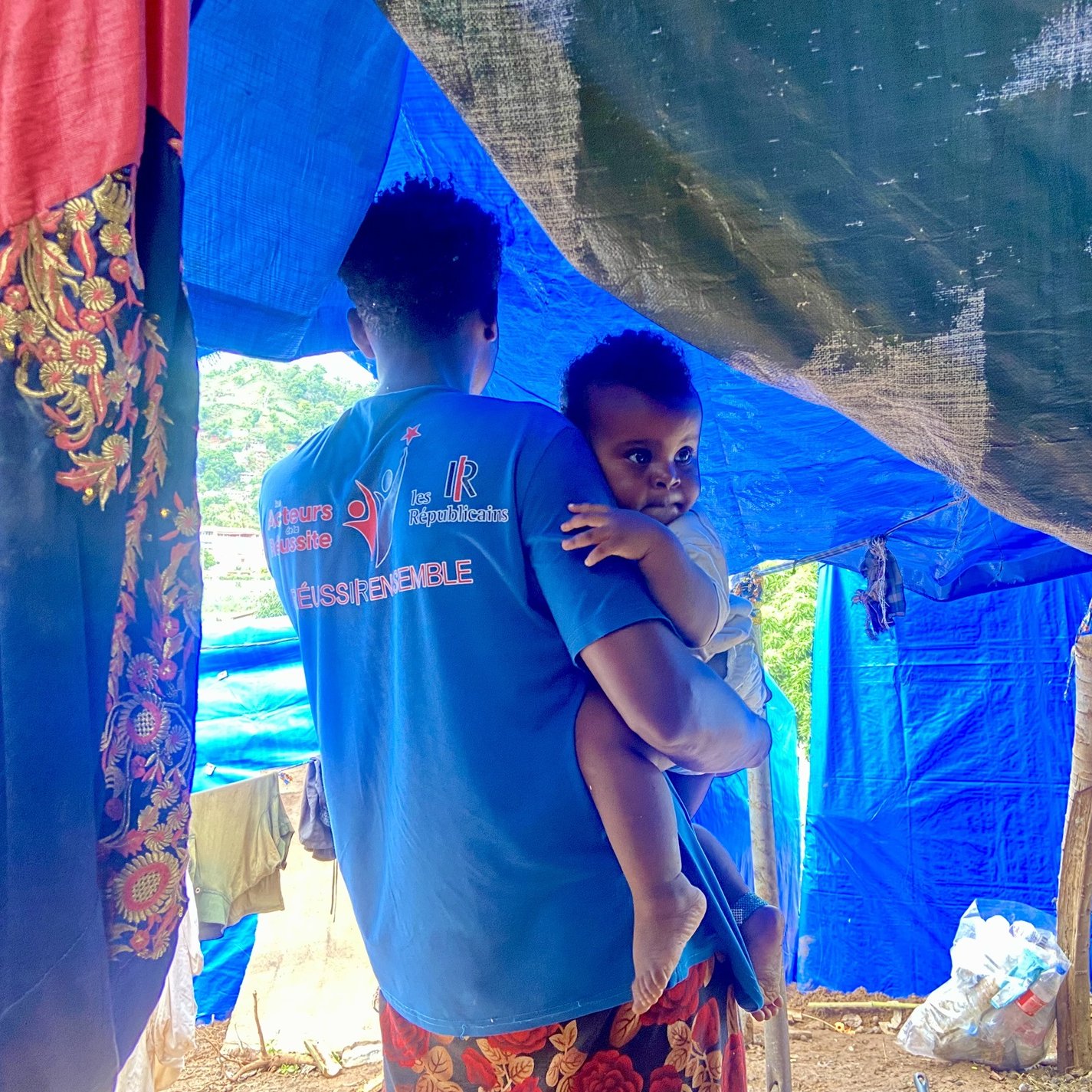 L'image montre un homme portant un enfant dans ses bras. Ils se trouvent sous une tente improvisée faite de tissus aux couleurs vives comme le bleu et le rouge. Le décor semble rural, avec des éléments naturels en arrière-plan. L'homme a un t-shirt bleu avec des inscriptions, et l'enfant, qui regarde légèrement en arrière, semble être de très jeune âge. La scène évoque un moment de tendresse et de protection dans un environnement simple.