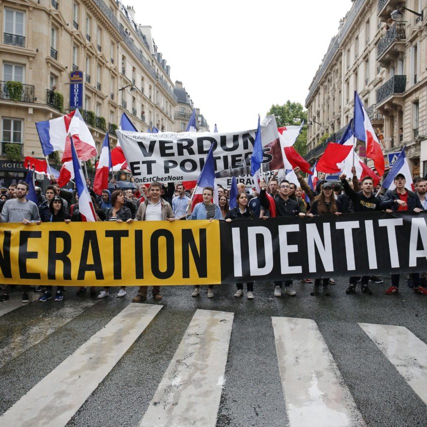 L'image montre une manifestation dans une rue urbaine. Un groupe de personnes tient une grande banderole noire avec le texte "GENERATION IDENTITAIRE" en lettres jaunes bien visibles. Elles brandissent également de nombreux drapeaux tricolores français, qui flottent au-dessus d'elles. En arrière-plan, on aperçoit une autre bannière avec les mots "VERDUN ET POITERS", indiquant probablement des messages ou des revendications relatives à ces lieux. Les manifestants semblent unis et déterminés, marchant ensemble sur le passage piéton, tandis que des bâtiments typiques de la ville se dressent à leurs côtés. L'ambiance générale est celle d'une démonstration publique, avec une forte présence de symboles nationaux.