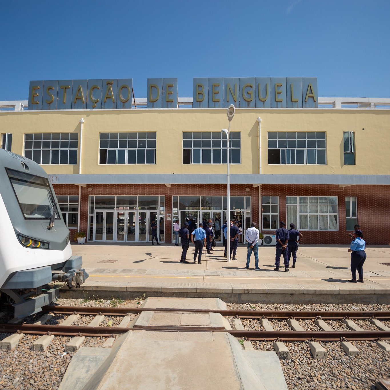 The image shows the exterior of a train station labeled "ESTAÇÃO DE BENGUELA." The building has a modern architectural design with large glass windows and a clean facade. In front of the station, a group of people, likely staff or passengers, are gathered. To the left, there is a sleek train parked on the tracks, showcasing a contemporary design. The scene is bright, indicating a clear blue sky. The overall setting suggests a bustling transportation hub.