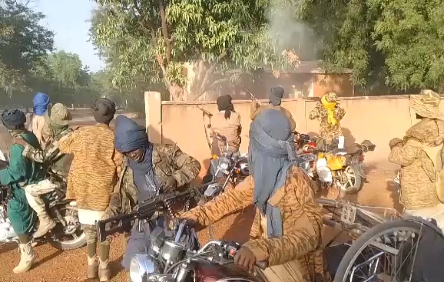 L'image montre un groupe de personnes sur des motos, portant des vêtements qui masquent leur visage et leur tête, probablement pour des raisons de camouflage ou de protection. Ils semblent être dans un environnement extérieur, avec des arbres et un bâtiment en arrière-plan. Il y a de la fumée qui s'échappe, indiquant peut-être une situation de tension ou d'activité intense. Certains individus tiennent des armes, ajoutant une atmosphère de gravité à la scène. Les motos sont variées et leur position suggère qu'ils sont en mouvement ou prêts à partir.