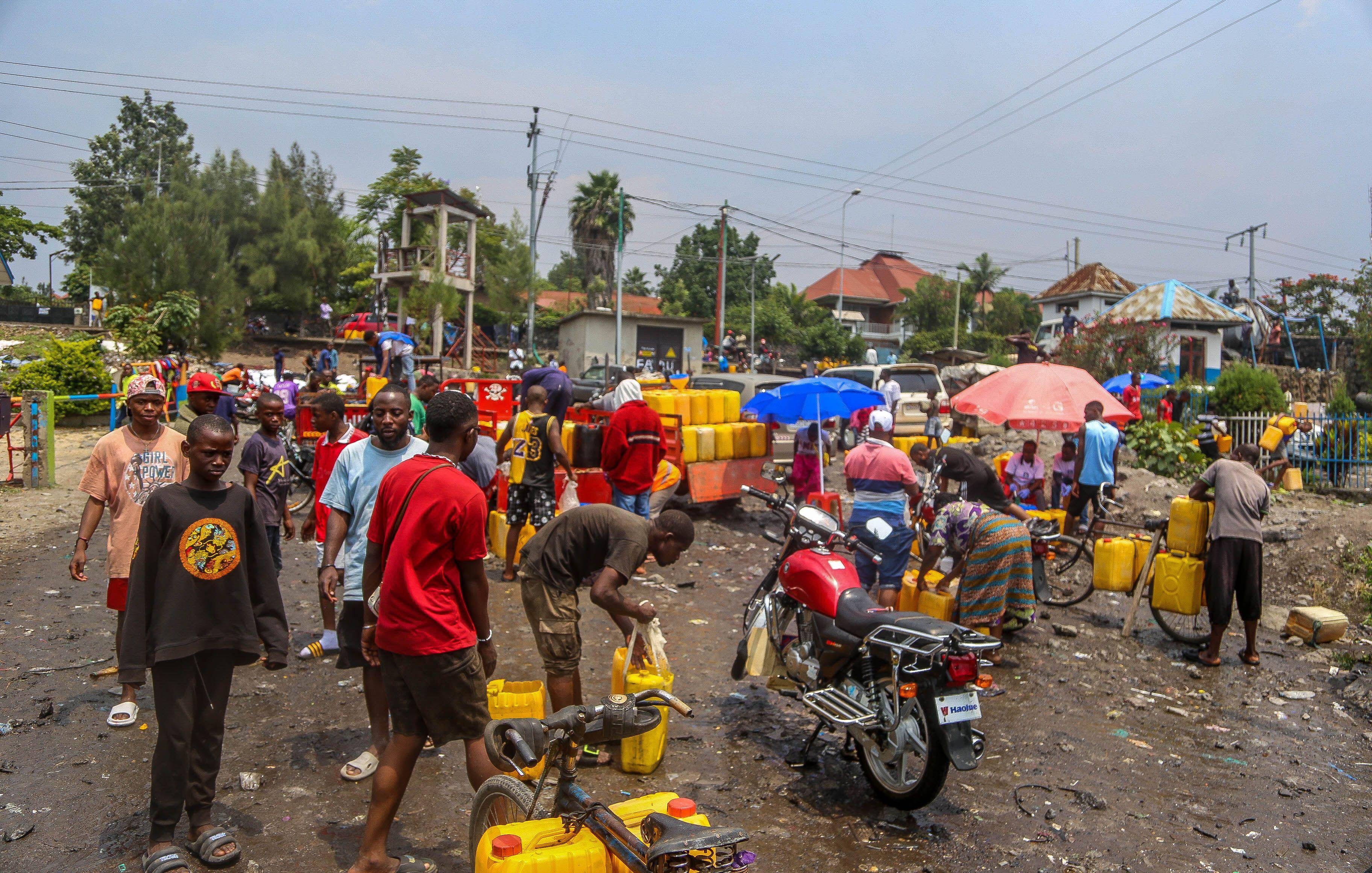 L'image représente une scène animée dans un quartier urbain. On peut imaginer un marché ou un point de distribution d'eau, où de nombreuses personnes sont rassemblées. Des hommes et des femmes, de divers âges, s'affairent autour de grands récipients jaunes qu'ils remplissent d'eau. Le sol semble en désordre, avec des déchets visibles, ajoutant une impression de négligence dans l'environnement. Des parapluies colorés sont déployés, fournissant un peu d'ombre sous un ciel légèrement nuageux. Au fond, on peut apercevoir des bâtiments et des arbres, tandis qu'un motard fait la queue avec sa moto, indiquant une activité routière environnante. L'atmosphère est dynamique, avec une forte interaction entre les gens.
