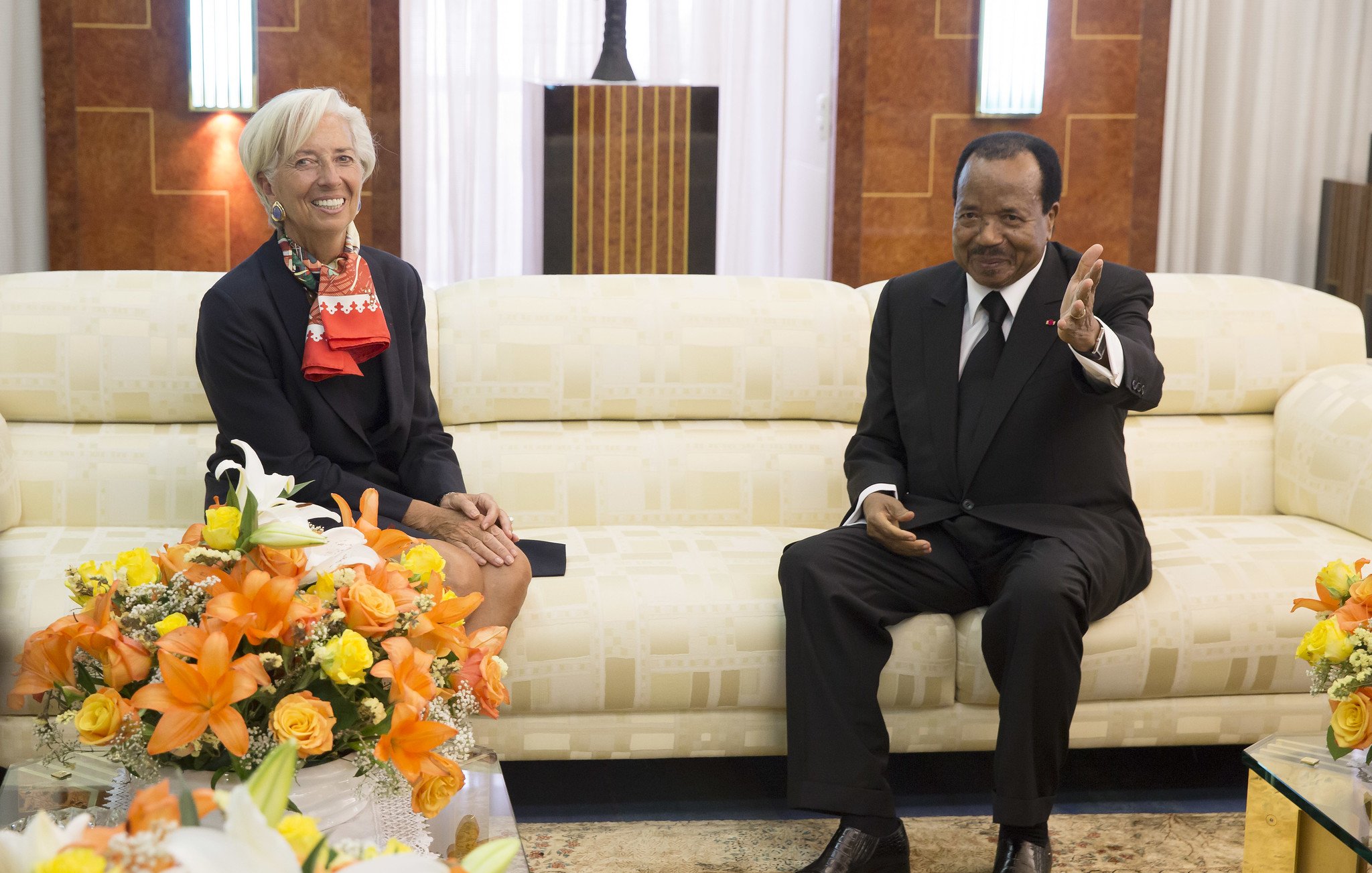 L'image montre deux personnes assises dans un salon élégant. À gauche, une femme aux cheveux courts et blonds, portant un blazer noir avec une écharpe rouge, sourit et semble détendue. À droite, un homme en costume noir et chemise blanche, assis sur un canapé crème, lève la main en signe de bienvenue ou pour parler. Ils sont entourés de bouquets de fleurs colorées, dominés par des teintes orange et jaune, qui ajoutent une touche de chaleur à l'environnement. Le décor est raffiné, avec des murs en bois et un éclairage doux, créant une ambiance conviviale.
