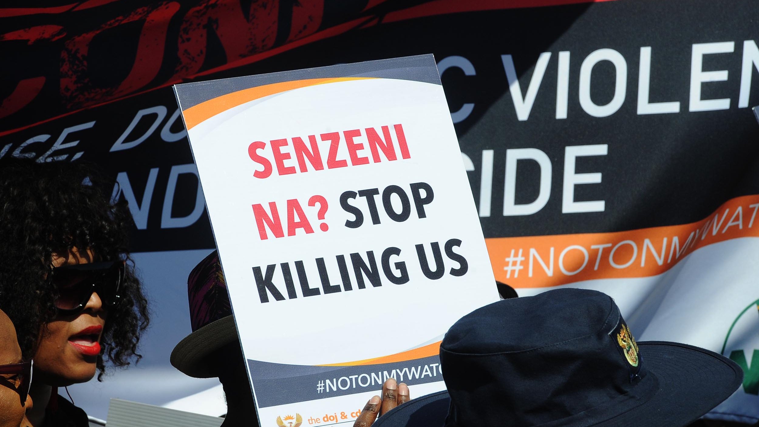 L'image montre un groupe de manifestants rassemblés pour une cause importante. Au premier plan, une personne tient une pancarte avec le slogan « SENZENI NA? STOP KILLING US ». Ce message manifeste un appel urgent à mettre fin à la violence et aux meurtres. En arrière-plan, il y a d'autres bannières et slogans, suggérant une atmosphère de protestation. Les personnes présentes portent des vêtements variés, y compris des chapeaux, et semblent engagées dans la cause qu'elles défendent. L'ambiance générale est celle de la solidarité et de la lutte pour la justice.