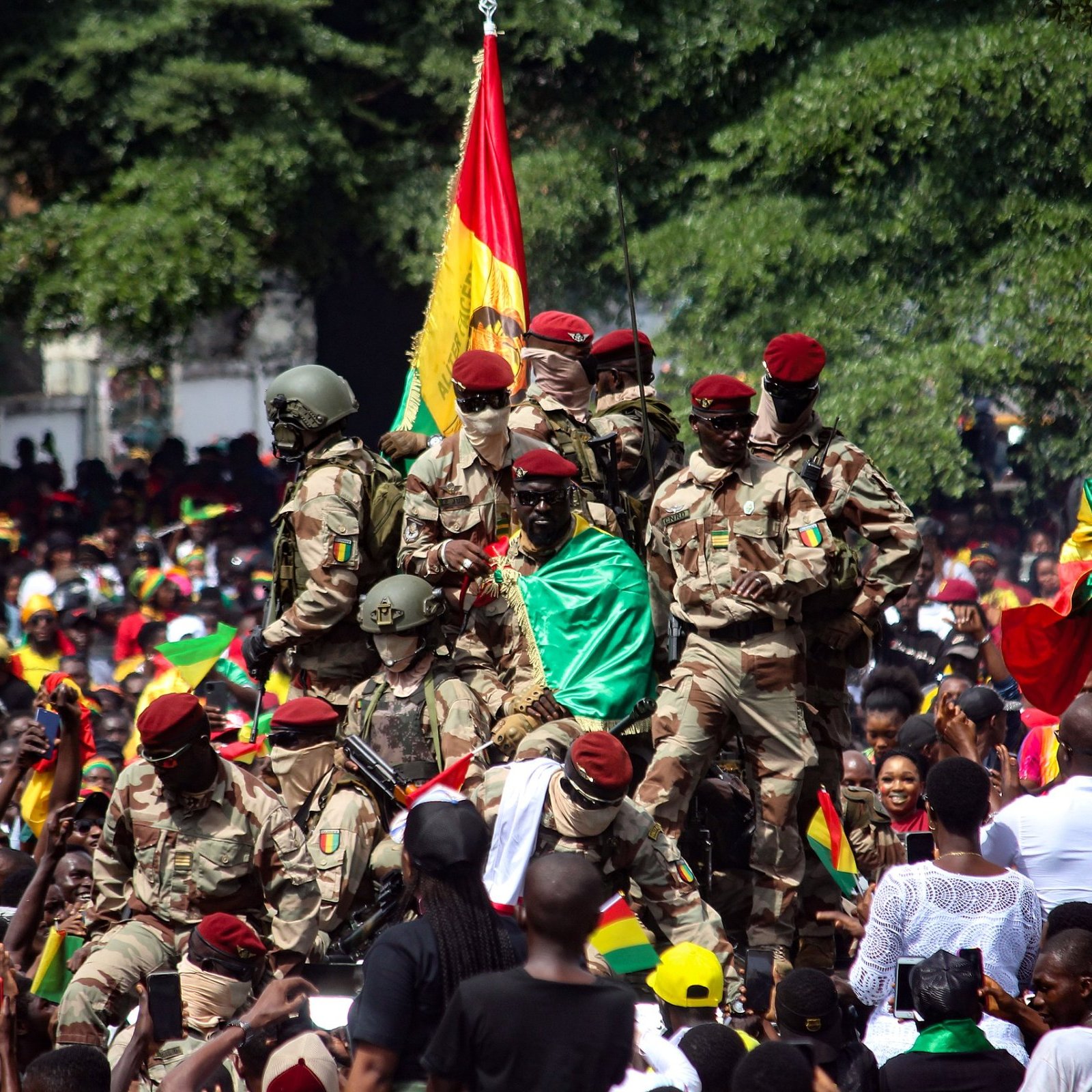 L'image montre une scène de célébration animée. Au premier plan, un groupe de soldats en uniforme militaire se tient debout, certains brandissant un drapeau aux couleurs vives de la Guinée, rouge, jaune et vert. Ils semblent être au centre d'une foule massive, qui est remplie de personnes qui portent également des vêtements aux couleurs du drapeau. L'ambiance est festive et euphorique, avec des visages souriants et levés vers les soldats. En arrière-plan, on aperçoit des arbres et des bâtiments, créant un cadre urbain. L'énergie collective de la foule et la présence militaire suggèrent un événement significatif, probablement une célébration ou une manifestation patriotique.