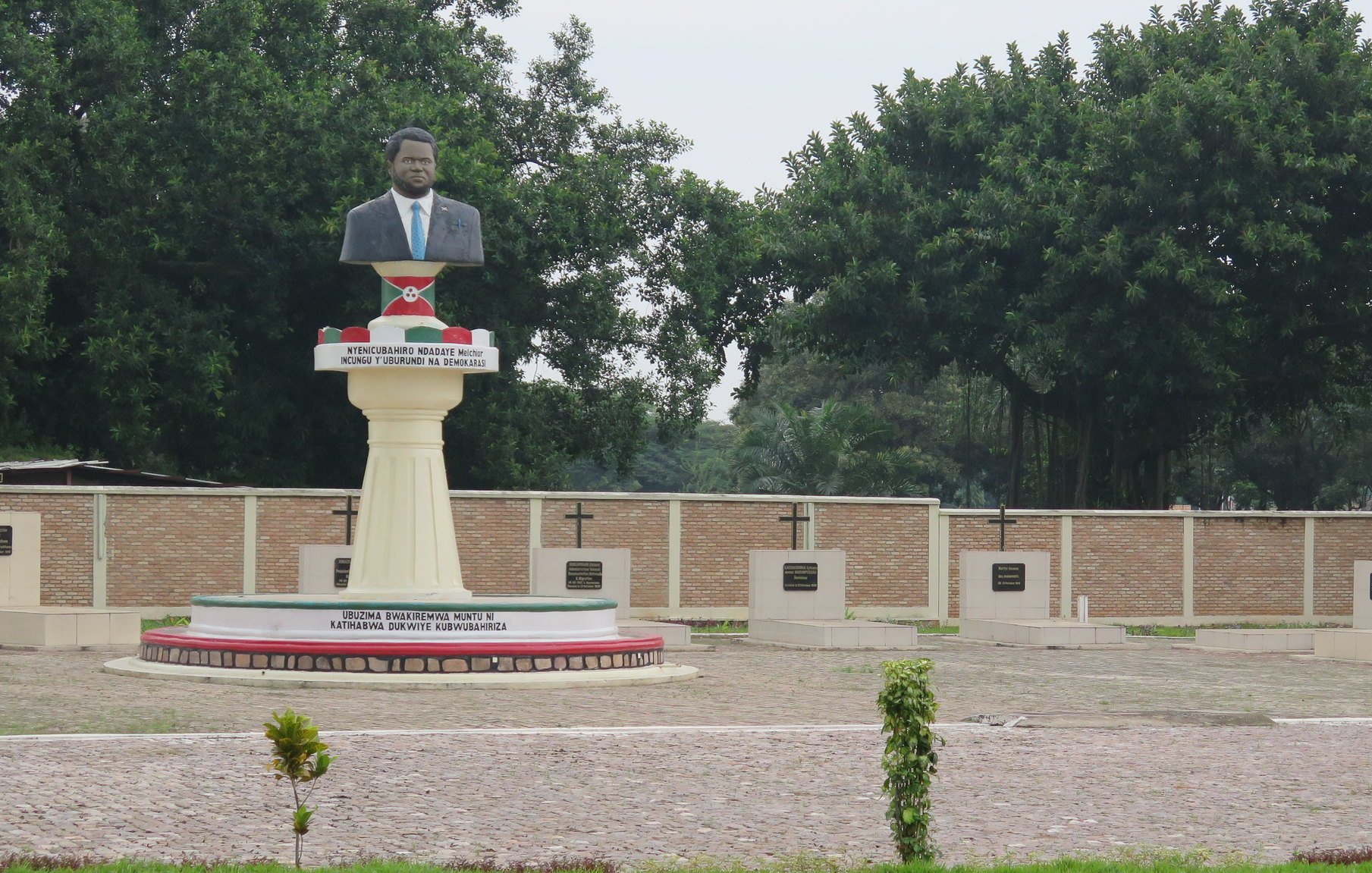 Cette image montre une statue en l'honneur d'une personnalité importante, probablement un leader ou un héros national. La statue est placée sur un socle élevé et représente un homme, vêtu d'un costume. En arrière-plan, on peut voir un mur avec plusieurs croix, qui semblent être des stèles commémoratives. Le site est entouré de verdure, avec des arbres touffus qui apportent une atmosphère paisible. Le sol est en pavé, et des plantes décoratives sont présentes, ajoutant une touche de nature à cet espace de mémoire.
