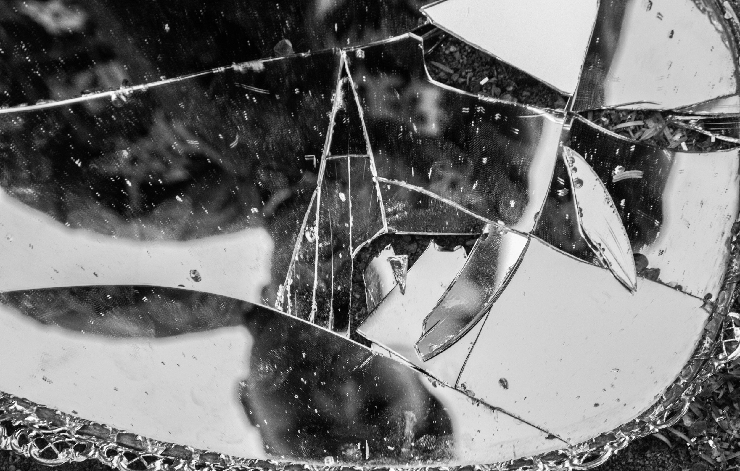 L'image présente un miroir brisé, dont les morceaux reflètent de manière fragmentée l'environnement. Les éclats de verre montrent des bords abrupts et irréguliers, créant une texture variée. La lumière joue sur la surface des fragments, produisant des éclats scintillants. La composition monochrome en noir et blanc accentue les contrastes entre les zones sombres et lumineuses, apportant une ambiance dramatique à l'ensemble. L'impression générale évoque une sensation de fragilité et de désordre, tout en incitant à la réflexion sur les thèmes de la rupture et de la beauté dans l'imparfait.