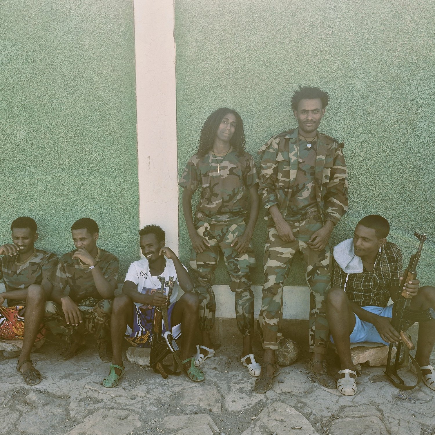 L'image montre un groupe de jeunes hommes, assis et debout, le long d'un mur vert. Les hommes portent des uniformes militaires camouflés pour certains, et d'autres sont habillés de manière plus décontractée. Ils semblent être dans un environnement extérieur, avec un sol en pierre. Quelques-unes des personnes tiennent des objets qui ressemblent à des sacs ou des équipements militaires. L'ambiance est tranquille, mais dégage une impression de camaraderie parmi le groupe.