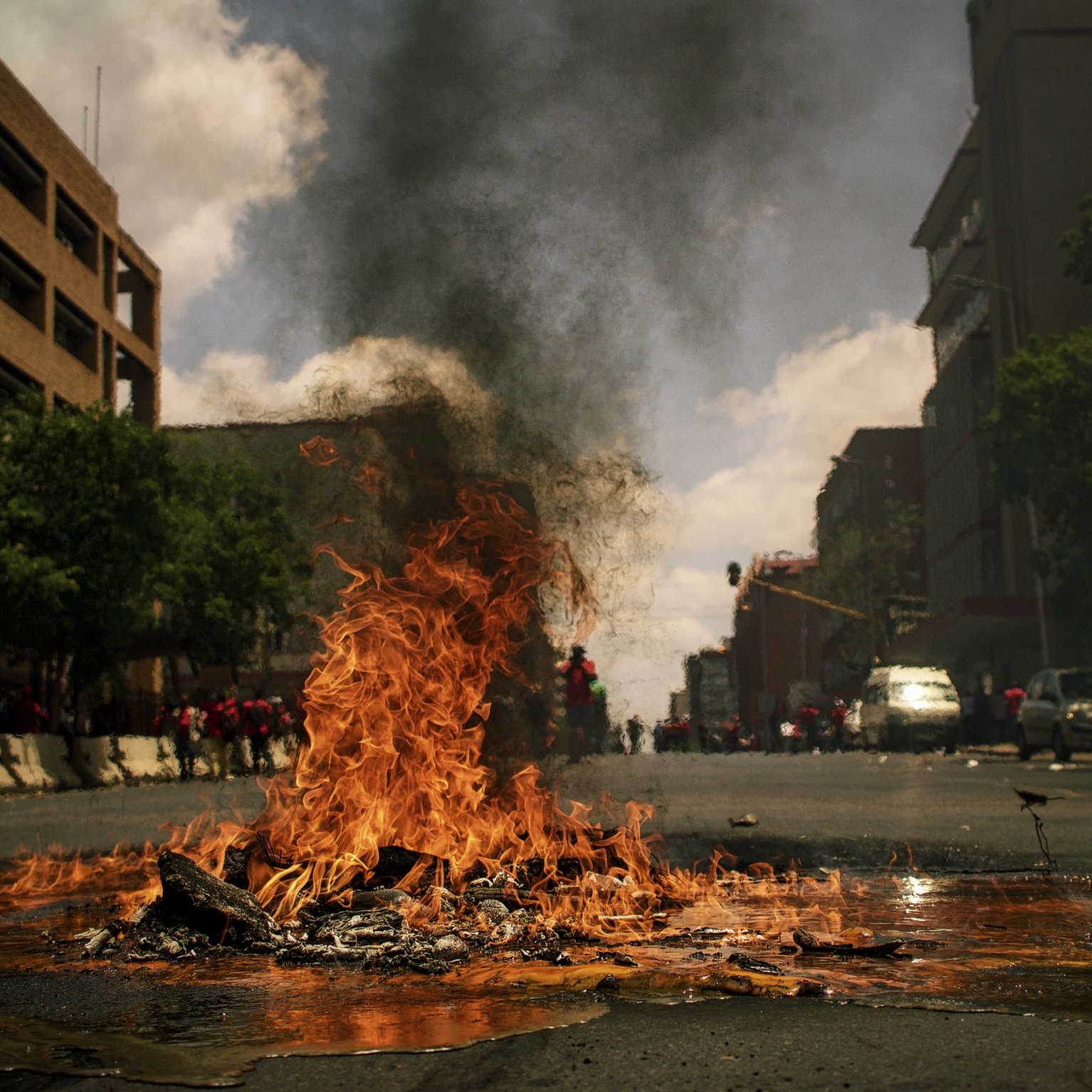 L'image montre une rue animée où un feu brûle au centre. Des flammes vives et tourbillonnantes s'élèvent de ce qui semble être des débris sur le sol. Une épaisse fumée noire s'échappe vers le ciel, obscurcissant partiellement la lumière du jour. On peut apercevoir des bâtiments sur les côtés de la rue, ainsi qu'un groupe de personnes au fond, créant une atmosphère tendue et dynamique. L'air est probablement chargé d'odeurs de brûlé, et le son du crépitement du feu se mêle à une ambiance vibrante de manifestation ou d'agitation.