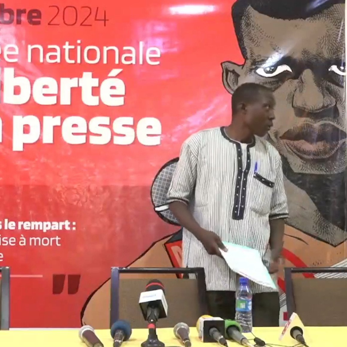 L'image montre un événement lié à la Journée nationale de la liberté de la presse, qui se déroule le 20 octobre 2024. En arrière-plan, un grand panneau rouge présente le thème de la journée : "Ne brisons pas le rempart : alerte sur la mise à mort du journalisme". Au premier plan, un homme debout s'apprête à s'adresser à l'audience. Il porte une chemise à rayures noires et blanches et se tient près d'une table équipée de plusieurs microphones et de bouteilles d'eau. L'ambiance est sérieuse et engageante, soulignant l'importance de la liberté d'expression.