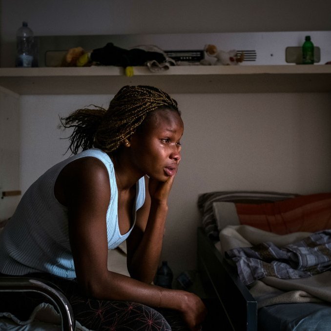 L'image dépeint une femme assise sur le bord d'un lit dans une pièce sobre. Elle a une coiffure tressée et porte un haut sans manches de couleur claire. Son expression est pensive, montrant une profonde réflexion ou peut-être de la tristesse. La lumière douce qui filtre à travers une fenêtre éclaire son visage, créant des ombres sur ses traits. La chambre semble être en désordre, avec des objets éparpillés et des draps non rangés. L'atmosphère de l'image évoque un sentiment de solitude et de contemplation.
