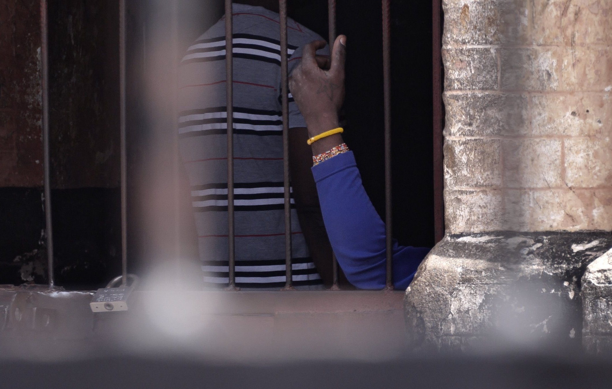 Cette image montre une scène à l'intérieur d'un espace confiné, avec des barreaux visibles au premier plan. On distingue des silhouettes humaines derrière les barreaux. Une main, portant un bracelet, s'élève légèrement, témoignant d'un geste de communication ou de contact. Les murs autour des barreaux semblent anciens et en pierre, ajoutant un élément d'authenticité à l'environnement. L'éclairage suggère que la scène se déroule à l'extérieur, avec une lumière naturelle qui éclaire partiellement l'espace.