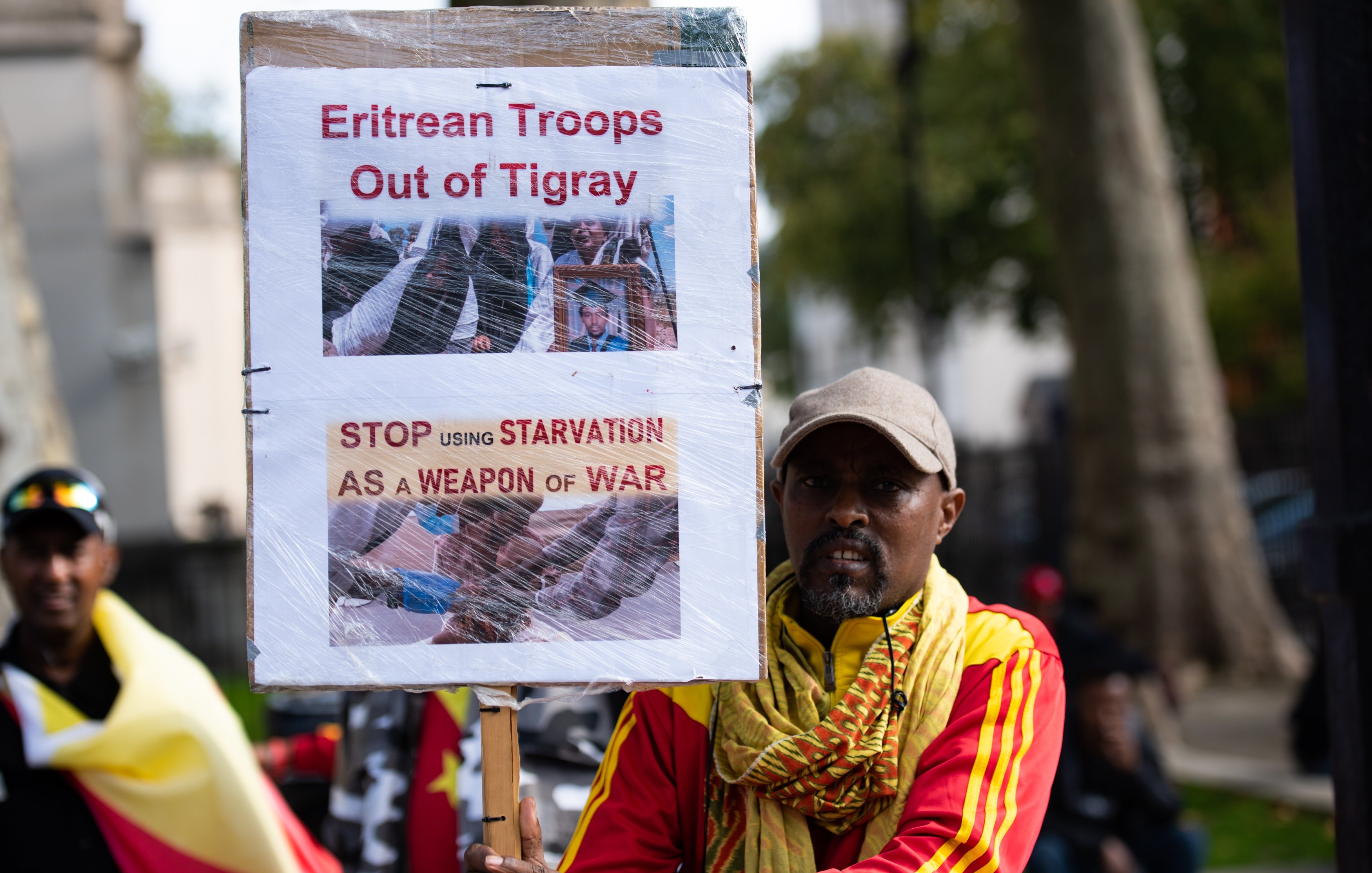 L'image montre un homme qui tient une pancarte haute, sur laquelle sont affichées des images et des textes en anglais. La pancarte exprime des revendications concernant le retrait des troupes érythréennes de la région du Tigré, en Éthiopie. Les mots en gras, "Eritrean Troops Out of Tigray" et "STOP USING STARVATION AS A WEAPON OF WAR", soulignent un appel urgent pour mettre fin à la souffrance causée par la guerre. L'homme porte un chapeau gris et est vêtu d'une écharpe colorée, probablement aux couleurs de l'Éthiopie, ce qui suggère un engagement émotionnel et une participation à une manifestation. En arrière-plan, on peut apercevoir d'autres personnes qui semblent partager des préoccupations similaires. L'ambiance est chargée d'émotion et de détermination.