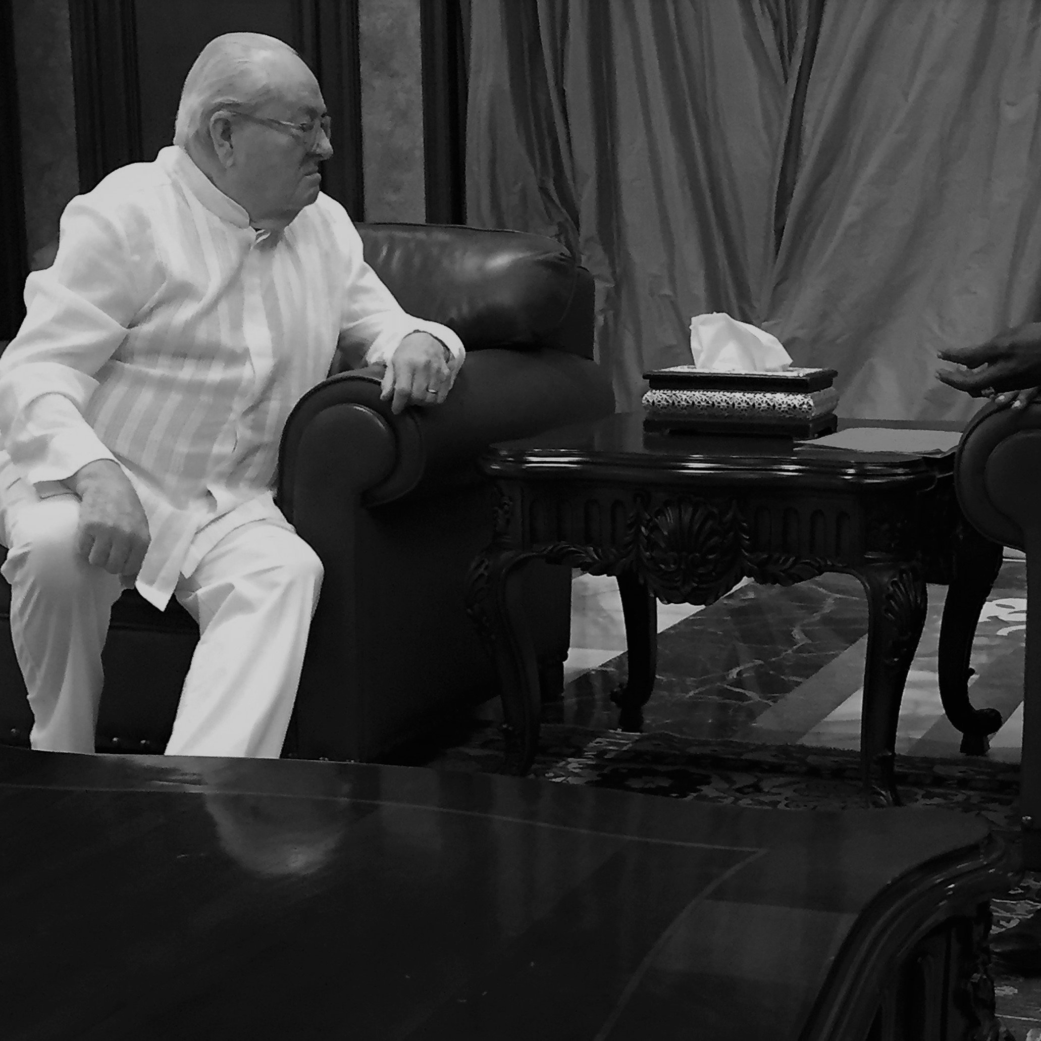 L'image représente une scène de discussion entre deux personnes assises l'une en face de l'autre dans un salon élégant. L'une des personnes porte un costume clair, tandis que l'autre est habillé d'un costume sombre. Ils sont assis sur des fauteuils en cuir, avec une table basse en bois ornée entre eux, où se trouve un objet décoratif. L'arrière-plan est composé de rideaux drapés et de murs en ton neutre, créant une atmosphère formelle et chaleureuse. L'éclairage est doux, ajoutant une sérénité à leur échange. On peut imaginer qu'ils discutent de manière engagée, leurs gestes accentuant l'intensité de la conversation.