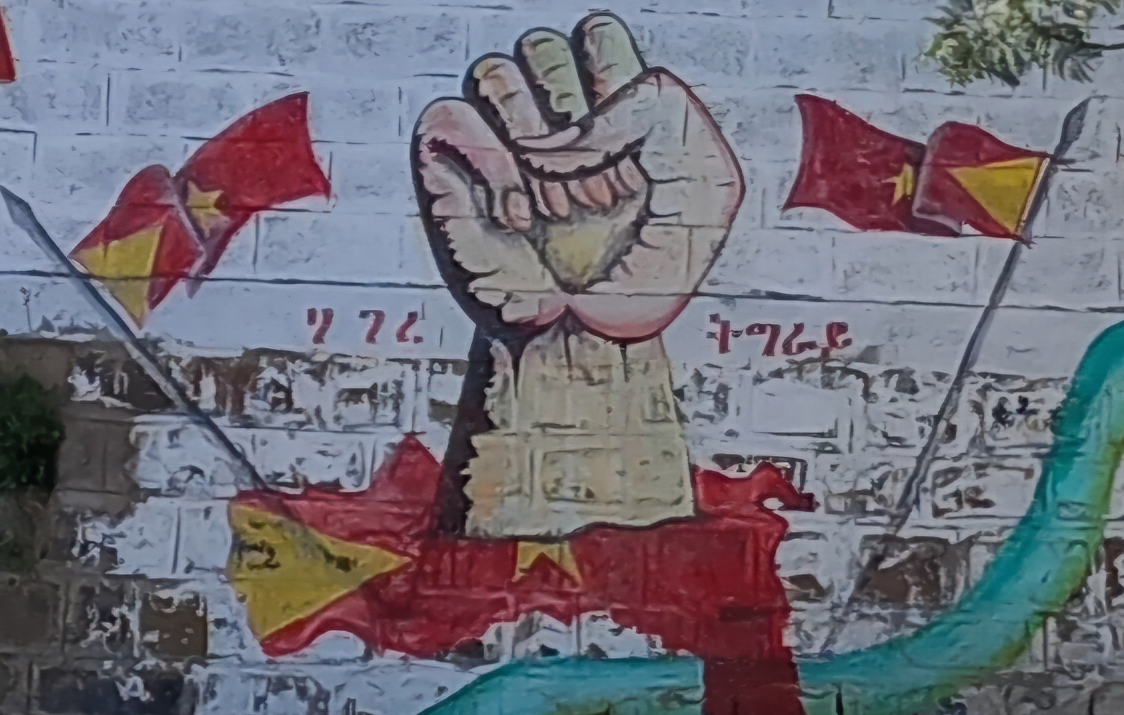 Sur le mur, on peut voir une grande fresque représentant un poing levé, symbole de résistance et de solidarité. Ce poing est peint dans des tons clairs, avec des détails qui soulignent sa force. Autour de lui, plusieurs drapeaux flottent, chacun arborant des motifs distincts en rouge et jaune, rappelant des symboles nationaux. En arrière-plan, on distingue des formes qui évoquent des paysages, créant une ambiance dynamique et engageante. L'ensemble transmet une forte impression de détermination et d'unité.