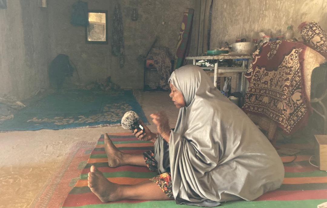 L'image représente une femme assise sur un tapis coloré dans un intérieur simple et rustique. Elle porte un vêtement ample et gris, qui couvre son corps. Sa posture est attentive, et elle tient un petit objet dans ses mains, probablement un instrument de musique ou une pièce d'artisanat. L'environnement autour d'elle est dépouillé, avec des murs en béton et quelques éléments de décoration, comme des tapis et des coussins. On peut sentir une atmosphère calme et intime, presque spirituelle. Des rayons de lumière pénètrent à travers une fenêtre, illuminant doucement la scène.