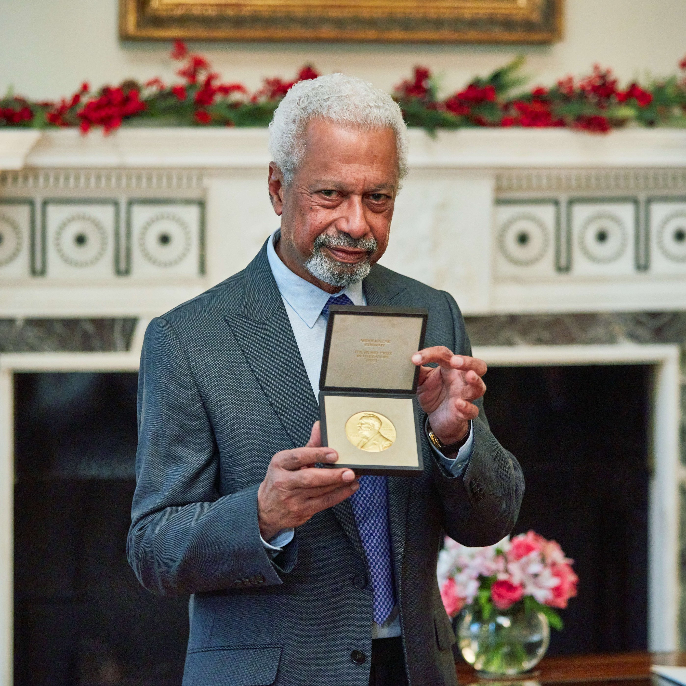 L'image montre un homme âgé, avec des cheveux poivre et sel et une barbe, qui porte un costume gris. Il se tient fièrement devant une cheminée décorée, tenant une médaille en or dans un écrin, qu'il montre à la caméra. En arrière-plan, on peut distinguer une décoration florale avec des fleurs disposées sur une table, créant une ambiance chaleureuse et élégante. La pièce est bien éclairée et a un air prestigieux, suggérant une occasion spéciale.