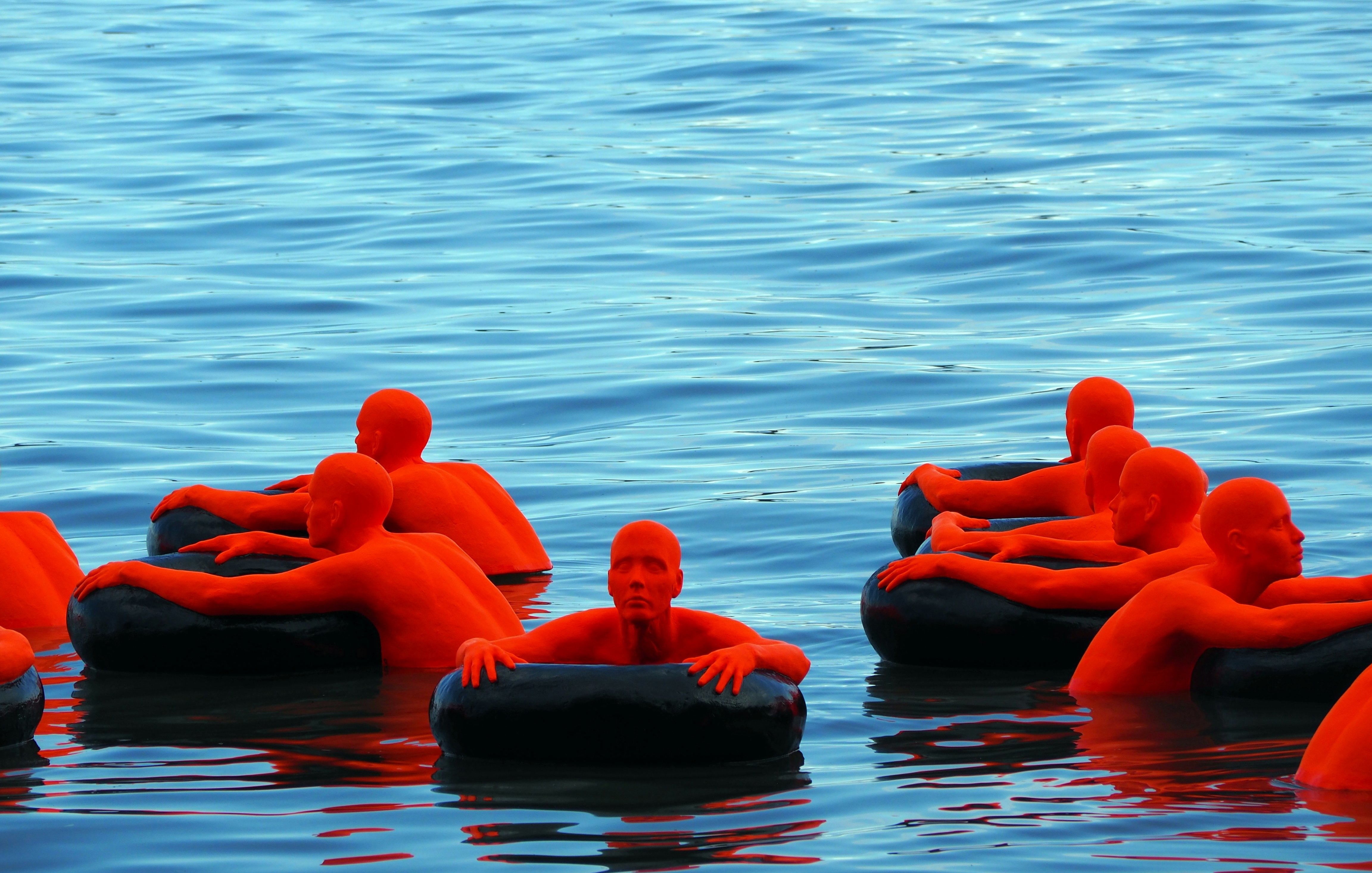 Cette image présente un groupe de sculptures humaines d'une couleur orange vif, flottant sur une surface d'eau calme. Chaque sculpture est assise ou allongée sur des anneaux flottants noirs, composant un contraste frappant avec le bleu azur de l'eau. Les sculptures ont des formes simplifiées, sans détails distincts de visage ou de vêtements, ce qui les rend abstraites. L'ensemble dégage une atmosphère à la fois sereine et intrigante, où la composition des éléments et les couleurs vives attirent l'attention.