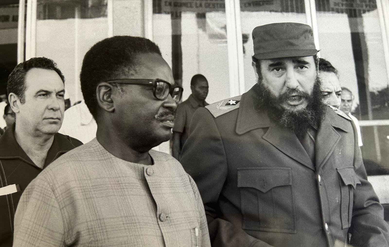 Cette image en noir et blanc montre trois hommes en discussion. Au premier plan, deux hommes sont particulièrement visibles. À droite, un homme avec une barbe fournie et un uniforme militaire, caractéristique de Fidel Castro, dialogue avec un homme portant des lunettes et un costume clair. Ce dernier semble attentif à la conversation. En arrière-plan, un troisième homme observe la scène. L'environnement est urbain, avec des bâtiments à vitre en arrière-plan. L'atmosphère paraît sérieuse et engagée, suggérant une rencontre diplomatique. Les expressions des visages laissent transparaître une certaine gravité.