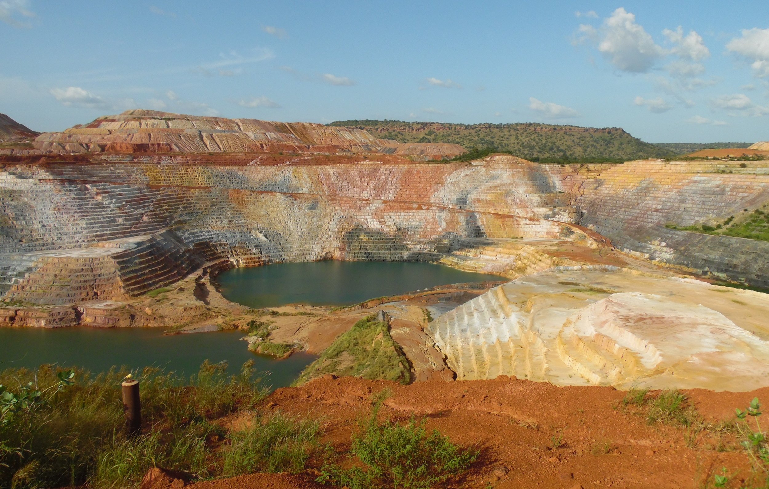 L'image montre une carrière de minerai, un paysage dramatique où les strates de terre créent des dégradés de couleurs allant du rouge au jaune et au blanc. Les parois de la carrière sont escarpées, formant des niveaux bien distincts. Au centre, un plan d'eau bleu émerge, contrastant avec les teintes terreuses et les paysages environnants. La végétation se fait rare sur les bords, mais quelques petites herbes vertes remontent le long des pentes. Le ciel est clair avec quelques nuages épars, ajoutant une lumière douce à la scène. Cette vue évoque à la fois l'exploitation humaine et la beauté naturelle brute.