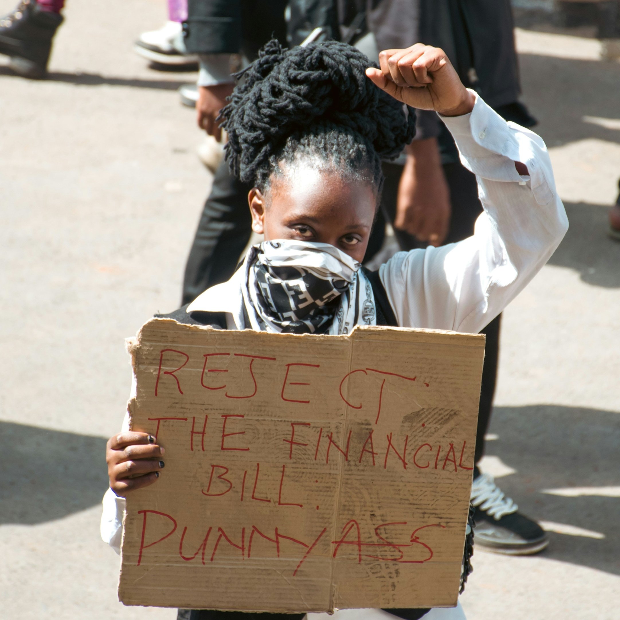 The image depicts a protester holding a cardboard sign that reads "REJECT THE FINANCIAL BILL PUNNYASS." The individual is wearing a white shirt and has their hair styled in large, coiled forms. They are raising a fist in a gesture of defiance or solidarity, and are also wearing a mask over their face. The background includes other people and a busy street scene, suggesting a gathering or demonstration.
