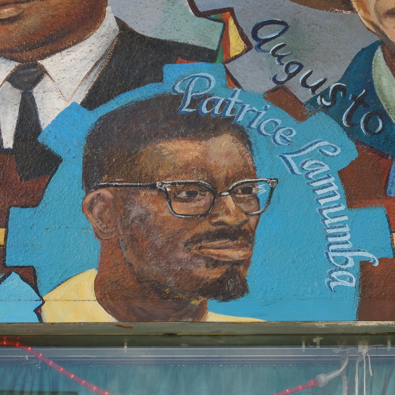 The image features a mural that includes a portrait of a man with glasses and a beard, identified as Patrice Lumumba. He is depicted in a colorful artistic style, wearing a yellow shirt. The mural may also include other figures or elements, but this particular part focuses on Lumumba's likeness alongside his name, highlighting his significance. The background is vibrant, contributing to the overall visual impact of the mural.