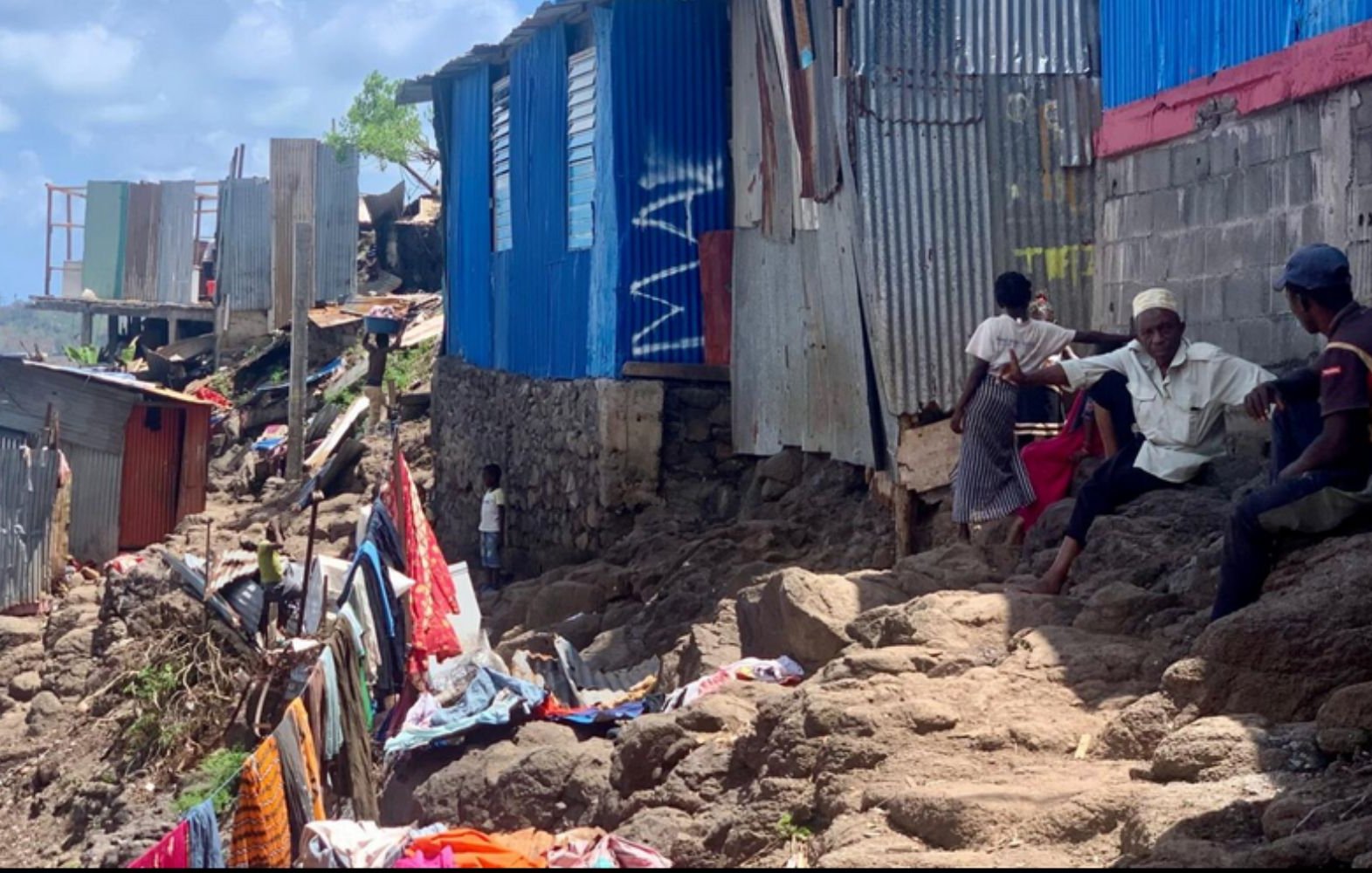 Cette image présente un paysage d'un quartier informel, caractérisé par des habitations en tôle et en béton, construite sur un terrain escarpé. Les maisons, souvent de différentes couleurs, semblent inachevées avec des parois en métal ondulé. Au premier plan, des personnes sont assises sur des rochers, discutant ou s'affairant. Le sol est accidenté, recouvert de débris et de vêtements épars, tandis qu'en arrière-plan, des structures réduites à des vestiges montrent l'impact de l'environnement sur ce lieu de vie. Le ciel est clair, laissant présager une journée ensoleillée. L'atmosphère générale évoque une communauté résiliente, malgré les difficultés et les conditions de vie précaires.