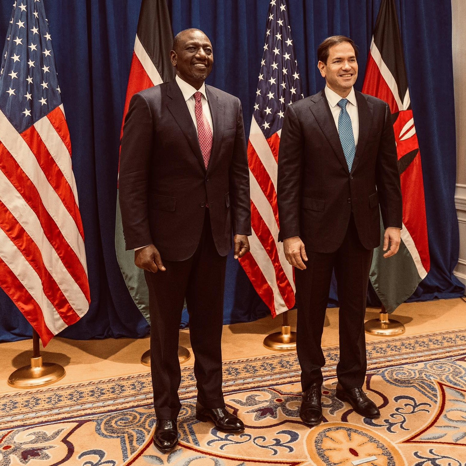 Sur cette image, nous voyons deux hommes debout côte à côte, souriants. À gauche, un homme portant un costume noir et une cravate rouge. Il a la peau foncée et une posture confiante. À droite, un homme avec un costume sombre et une cravate bleu clair. Il a la peau plutôt claire et un sourire engageant. Derrière eux, il y a trois drapeaux : deux des États-Unis, avec des étoiles blanches sur fond bleu et stripes rouges et blancs, et un drapeau du Kenya, qui a des bandes horizontales noires, rouges et vertes, avec un bouclier au centre. Le décor est élégant, avec un fond bleu et un tapis orné à leurs pieds. L'atmosphère semble formelle et amicale.