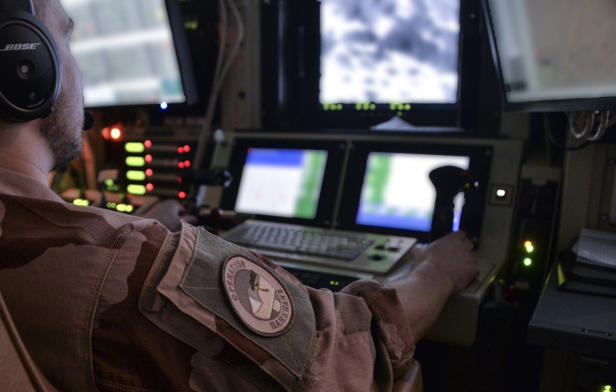 Dans cette image, on voit un homme assis devant un tableau de contrôle, portant un uniforme militaire. Il a un casque sur les oreilles et utilise des manettes et des boutons sur la console. Devant lui, un écran affiche des images en noir et blanc, suggérant qu'il surveille ou contrôle quelque chose à distance. Le décor est sombre, avec des lumières d'indication vertes et rouges autour de lui, ajoutant une atmosphère technologique et militaire à la scène. L'arrière-plan suggère un environnement de travail complexe, probablement dans un avion ou un centre de commandement.