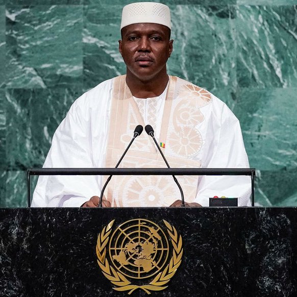 L'image montre un homme debout derrière un pupitre lors d'un discours. Il porte un costume traditionnel, probablement en blanc, avec un embellissement distinctif. Son teint est sombre et il a une expression sérieuse, indiquant qu'il est en train de s'adresser à un public. Le fond de l'image est constitué de panneaux de marbre vert, ce qui crée un contraste avec sa tenue. Le pupitre est orné du symbole des Nations Unies, représentant la mission internationale de paix et de coopération.