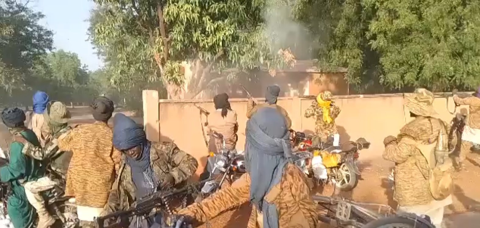 L'image montre un groupe de personnes sur des motos, portant des vêtements qui masquent leur visage et leur tête, probablement pour des raisons de camouflage ou de protection. Ils semblent être dans un environnement extérieur, avec des arbres et un bâtiment en arrière-plan. Il y a de la fumée qui s'échappe, indiquant peut-être une situation de tension ou d'activité intense. Certains individus tiennent des armes, ajoutant une atmosphère de gravité à la scène. Les motos sont variées et leur position suggère qu'ils sont en mouvement ou prêts à partir.