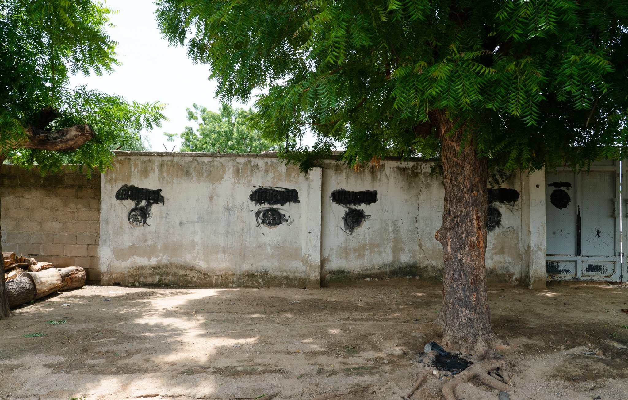 L'image présente un mur en béton aux couleurs neutres, probablement gris ou beige, partiellement recouvert par des ombres de feuillage provenant d'un grand arbre à proximité. Sur le mur, on peut voir des traces de peinture noire qui forment des motifs stylisés évoquant des yeux. Ces motifs sont positionnés de manière à créer une disposition particulière sur le mur. Au premier plan, on remarque également un tronc d'arbre robuste, avec des racines visibles qui émergent du sol sablonneux. L'environnement semble tranquille, avec une légère luminosité qui indique une journée ensoleillée.