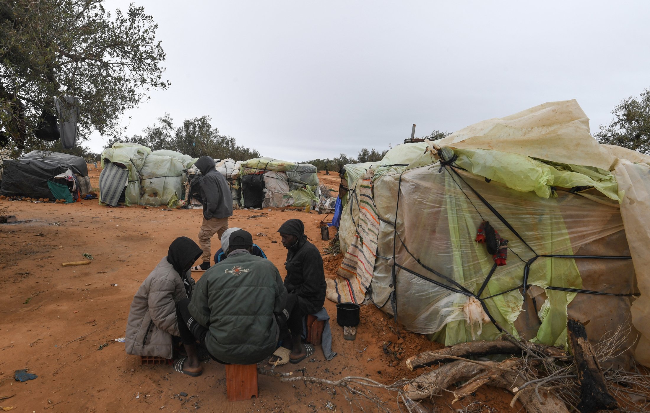 L'image montre un paysage d'abris précaires, construits avec des matériaux comme du plastique et des bâches, disposés sur un terrain sablonneux. Au premier plan, un groupe de personnes est assis en cercle, partagé entre une conversation ou une activité commune. Les abris semblent rudimentaires, entourés d'un environnement naturel avec quelques arbres épars. L'atmosphère est grise, suggérant un temps maussade. Skies sombres et nuageux donnent une impression de froid et de dureté.