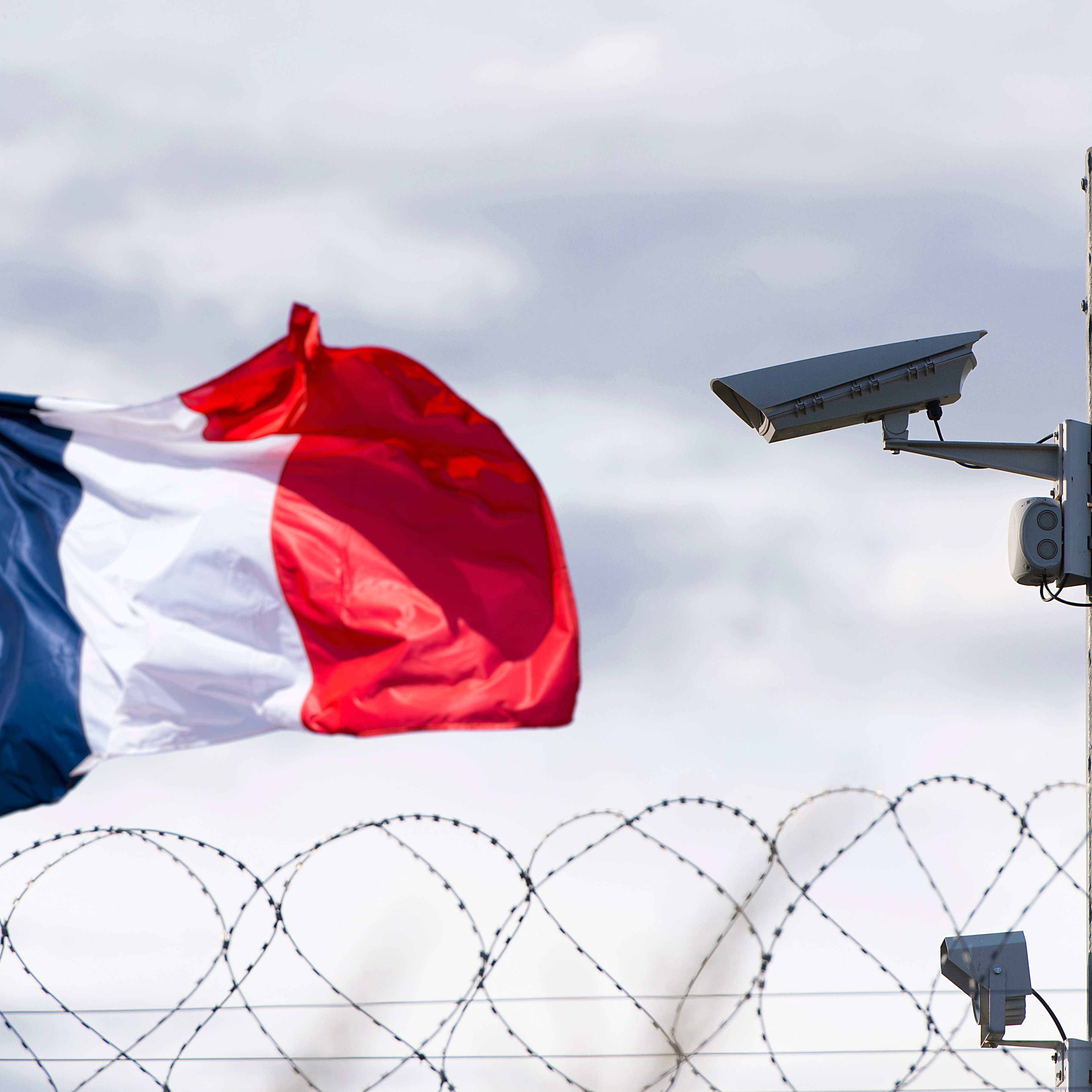 L'image montre un drapeau français vibrant, flottant au vent. À côté, on aperçoit une caméra de surveillance fixée sur un poteau. En arrière-plan, un fil de fer barbelé accentue l'idée de sécurité et de surveillance. Le ciel est nuageux, ajoutant une atmosphère à la scène, qui évoque un environnement protégé.