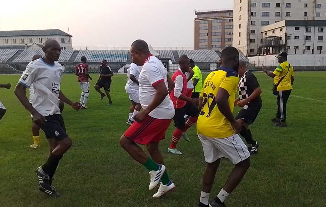 L'image représente un groupe de personnes jouant au football sur un terrain en herbe. Les joueurs portent des maillots de différentes couleurs, principalement blancs, rouges et jaunes. On peut voir des hommes en train de s'échauffer ou de danser, avec des mouvements dynamiques et joyeux. En arrière-plan, on aperçoit des gradins remplis, et des bâtiments se dressent autour du terrain. L'ambiance semble conviviale et énergique, avec un sentiment de camaraderie parmi les joueurs.