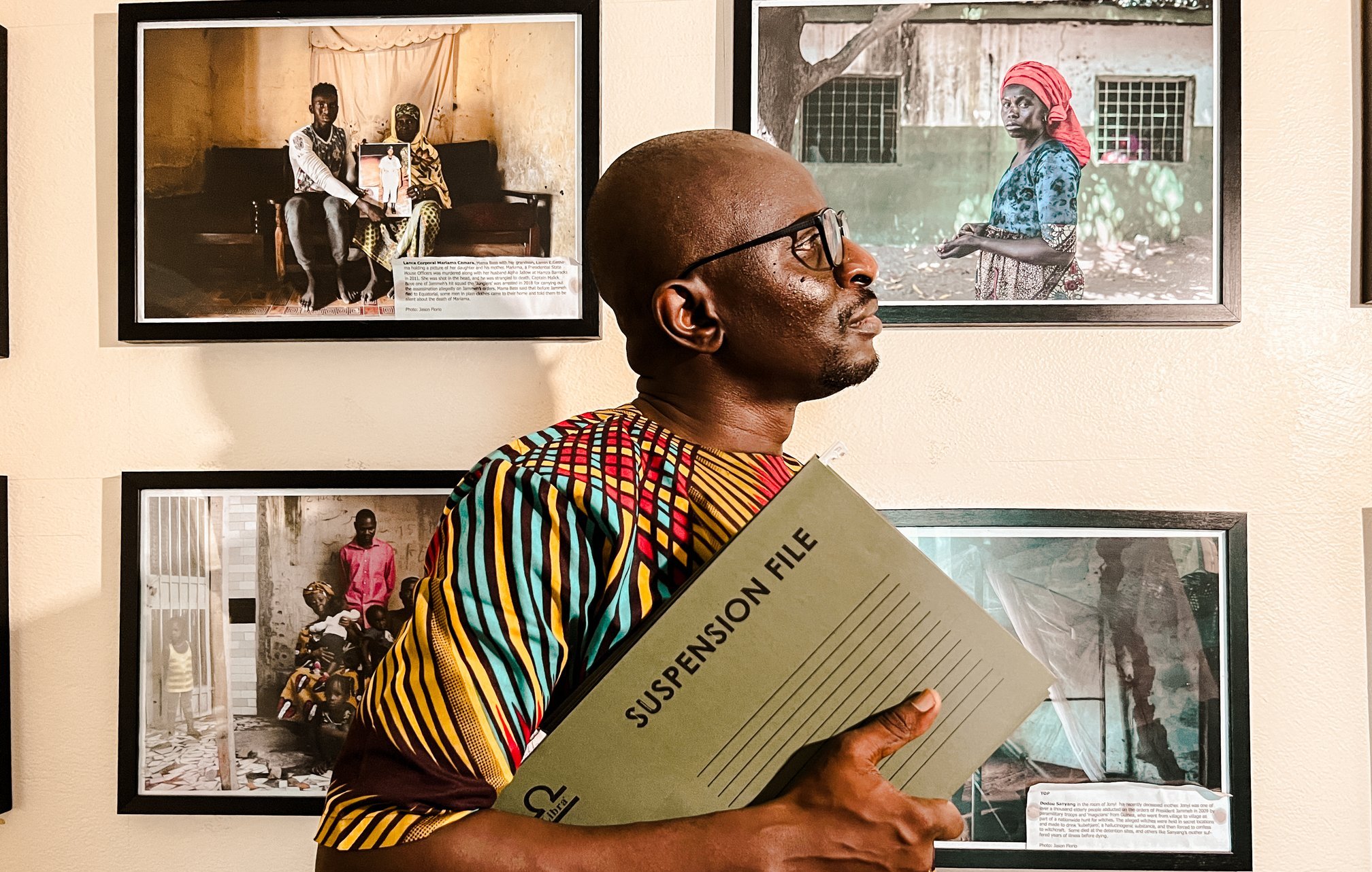 L'image montre un homme debout dans un espace d'exposition, entouré de plusieurs photographies accrochées au mur. Il porte une chemise colorée aux motifs vibrants, mêlant des teintes de jaune, rouge et vert. Dans ses bras, il tient un dossier intitulé "SUSPENSION FILE", ce qui suggère qu'il se prépare à discuter ou à présenter quelque chose d'important. L'homme se tourne légèrement sur le côté, regardant vers les photos, qui semblent raconter des histoires de vie, avec des portraits d'autres personnes. L'atmosphère de l'image est à la fois sérieuse et engagée, soulignant l'importance des histoires capturées dans ces œuvres visuelles.