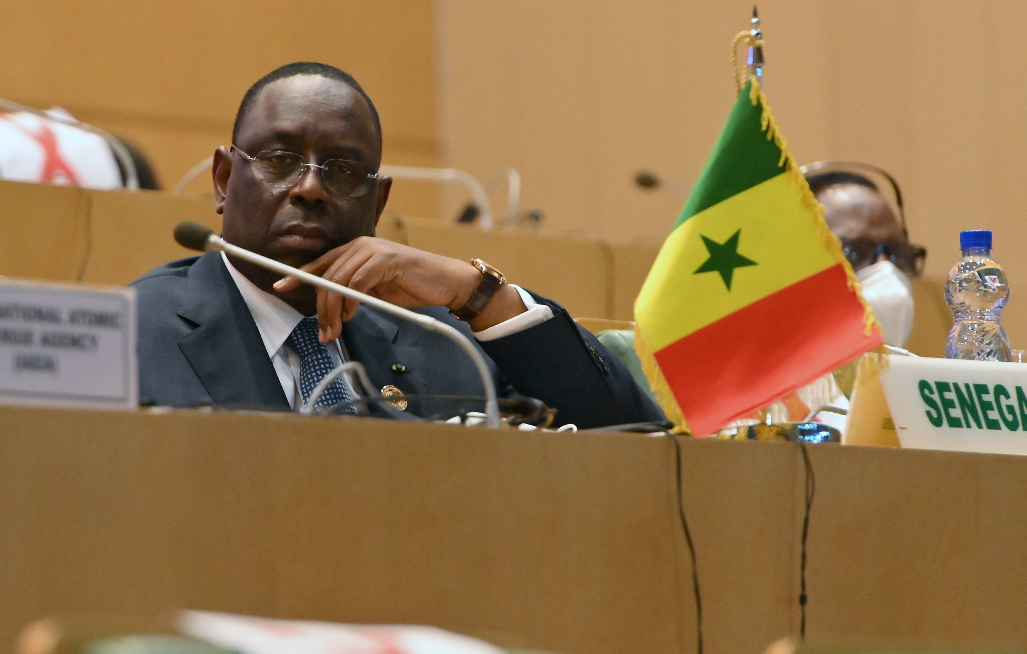 L'image montre un homme assis dans un cadre officiel, probablement lors d'une réunion ou d'une conférence. Il porte un costume sombre avec une chemise et une cravate, et affiche une expression pensive ou attentive. À sa droite, un petit drapeau du Sénégal est visible, avec les couleurs rouge, jaune et vert, ainsi que l'étoile verte au centre. On peut également apercevoir une bouteille d'eau à gauche et un badge ou un panneau indiquant "SENEGAL" devant lui. L'ambiance générale est sérieuse et concentrée, typique de discussions diplomatiques ou politiques.