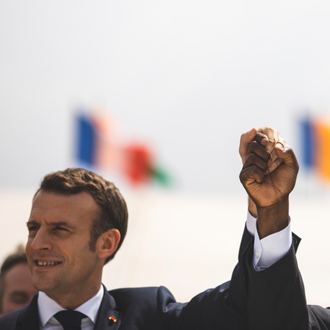 The image shows two men, one wearing a suit with a tie, raising their hands together in a gesture of solidarity or celebration. They are smiling and appear to be in a public setting. In the background, there are several flags visible, suggesting an international context or event. The atmosphere seems positive and hopeful.