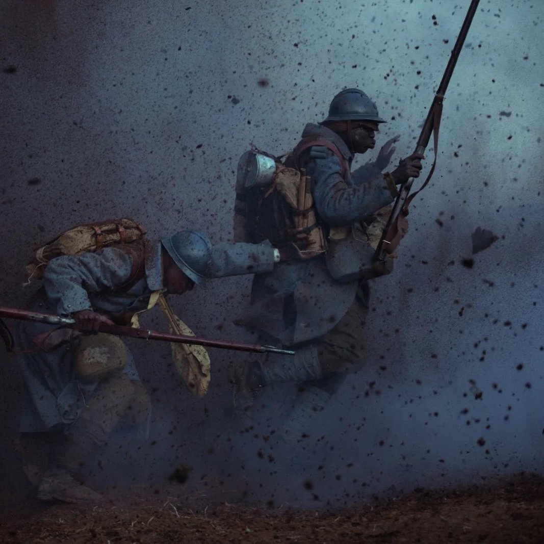 L'image dépeint une scène de guerre intense. Deux soldats, habillés de uniformes bleu ciel typiques de la Première Guerre mondiale, avancent avec détermination à travers un paysage chaotique. On ressent une ambiance de tension et de danger, accentuée par des éclats de poussière et de débris qui volent autour d'eux, comme si une explosion venait de se produire. Les soldats portent des sacs à dos lourds et sont armés de fusils, montrant leur préparation au combat. Leur position angulaire suggère un mouvement rapide, soulignant le courage et l'adrénaline de la situation. L'éclairage est sombre, créant une atmosphère pesante, presque apocalyptique.