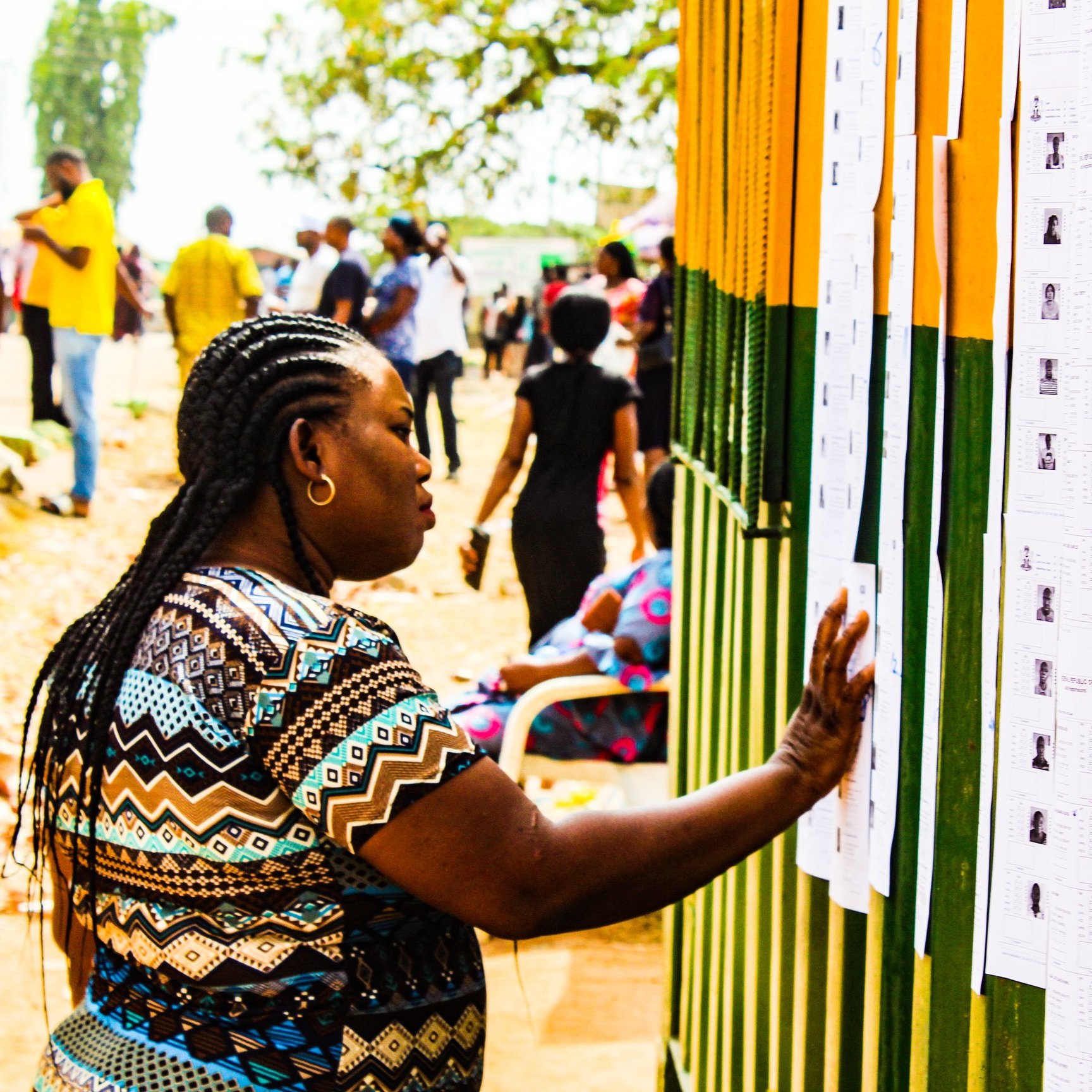 Dans cette image, on voit une scène animée en extérieur. Une femme, avec des tresses et portant un haut aux motifs colorés, se tient près d'un mur recouvert de plusieurs feuilles de papier. Elle passe sa main sur l'une de ces feuilles, qui semble contenir des informations, probablement des listes ou des résultats. En arrière-plan, de nombreuses autres personnes se déplacent, créant une atmosphère de rassemblement. Les couleurs vives du mur et la lumière du jour ajoutent une ambiance chaleureuse à la scène. Au sol, on peut apercevoir des feuilles mortes, ce qui suggère une saison automnale.