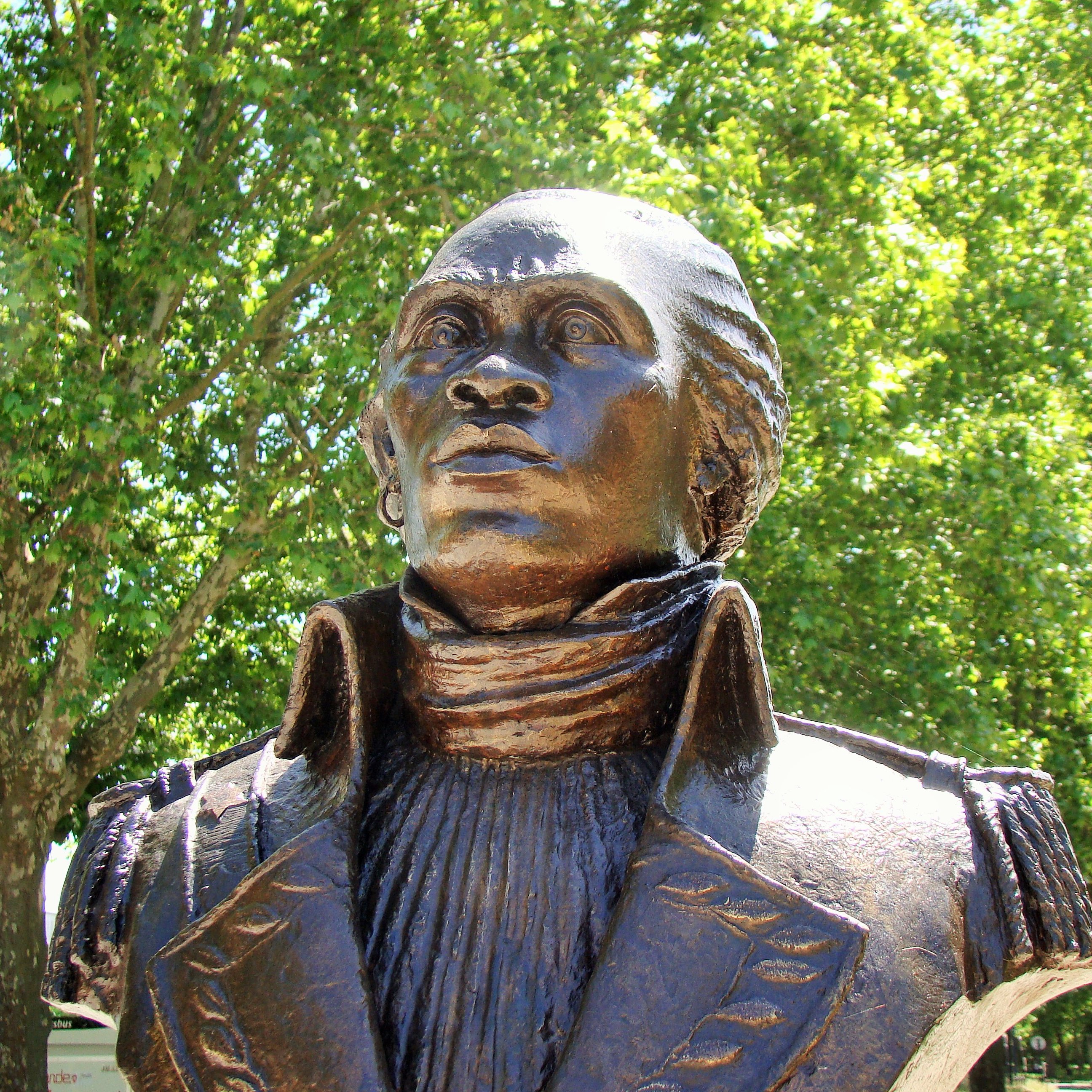 Voici la description de l'image : L'image montre une statue en bronze représentant un personnage en buste, probablement une figure historique. Le buste a un regard tourné vers le haut, suggérant une attitude déterminée ou réfléchie. Le personnage porte une veste à col haut, avec un plissé distinct sur le devant. Les détails du visage, bien que stylisés, montrent une expression forte, avec des traits bien définis. La statue est placée à l'extérieur, entourée d'arbres verdoyants, créant un cadre naturel agréable. La lumière du soleil illumine la statue, accentuant ses contours et sa texture.