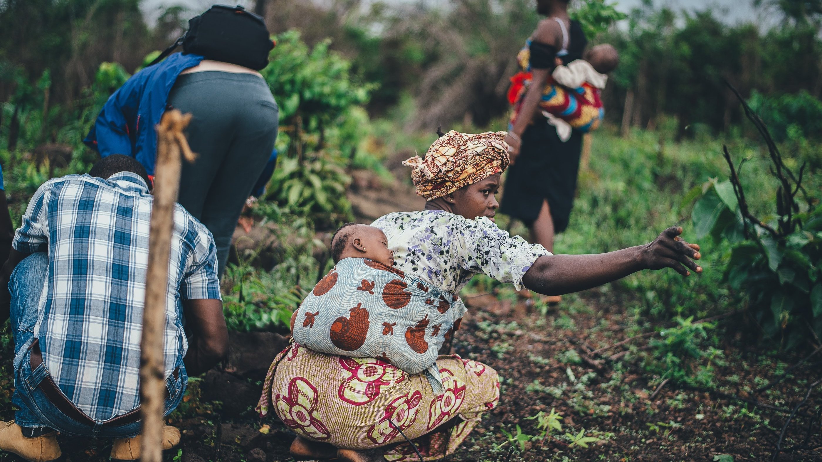L'image montre un groupe de personnes travaillant dans un champ verdoyant. Au premier plan, une femme porte un foulard coloré et un vêtement à motifs, avec un bébé enveloppé dans un porte-bébé sur son dos. Elle tend son bras pour indiquer quelque chose dans le champ. À l'arrière-plan, d'autres personnes, y compris une femme portant un enfant et un homme vêtu d'une chemise à carreaux, sont occupées à planter ou à récolter. L'atmosphère est dynamique et témoigne d'un effort collectif au milieu d'une nature luxuriante. On peut sentir une ambiance de travail, de solidarité et de connexion avec la terre.