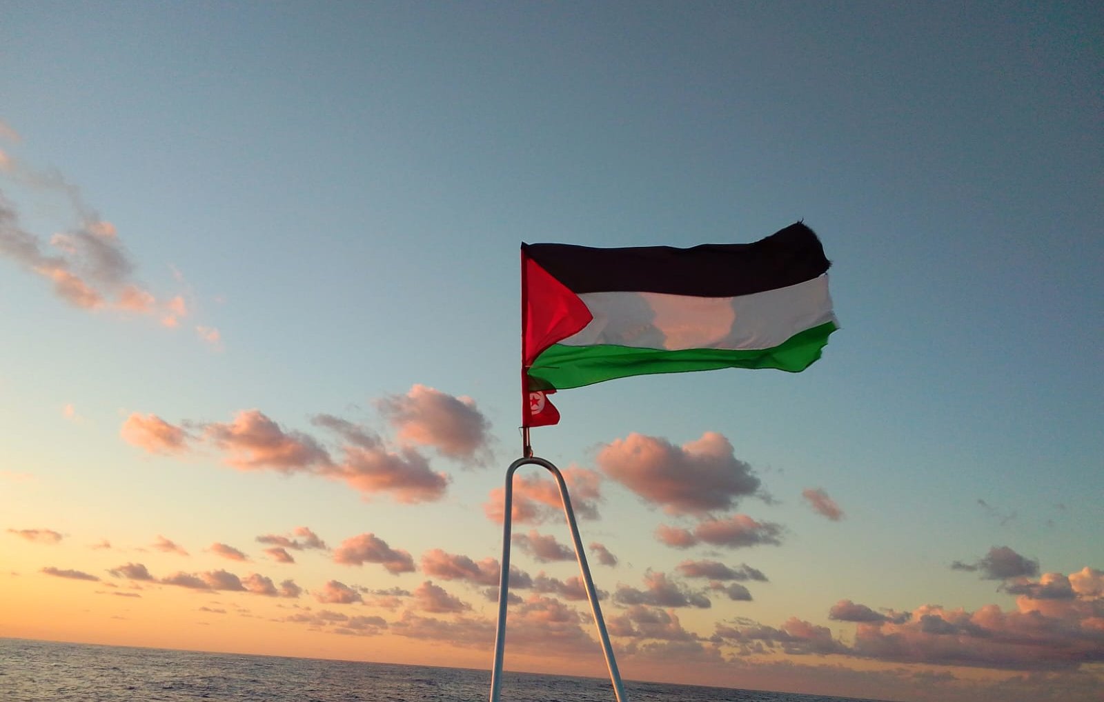 The image depicts a flag flying on a boat against a backdrop of a colorful sky during sunset. The flag features three horizontal stripes: black on the top, white in the middle, and green on the bottom, with a red triangle on the left side. The ocean can be seen below, and there are clouds in the sky that are illuminated by the setting sun, creating a serene atmosphere.