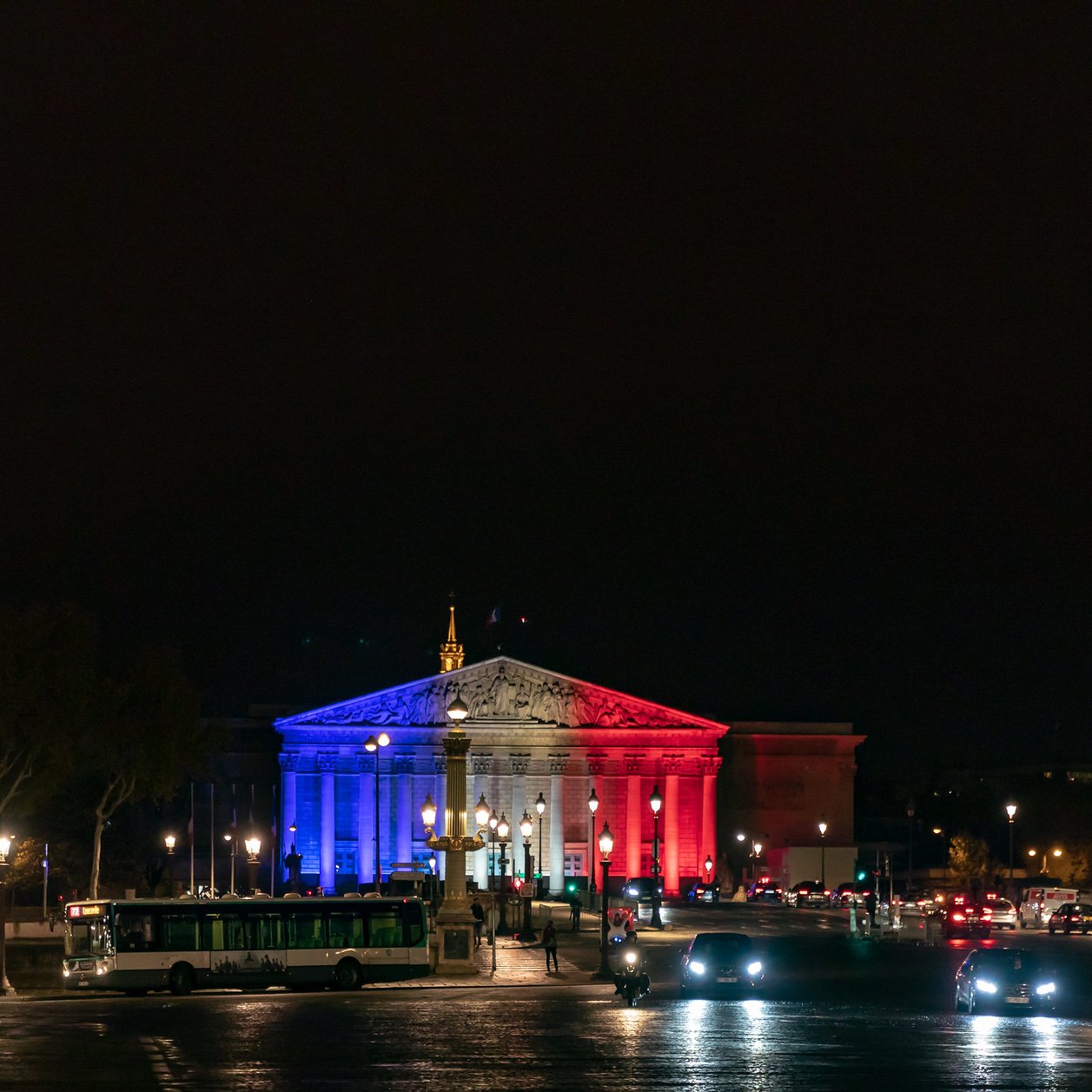 Imaginez un bâtiment majestueux illuminé par des lumières tricolores. La façade est peinte de bleu, de blanc et de rouge, évoquant les couleurs du drapeau français. Le ciel nocturne est sombre, créant un contraste saisissant avec les lumières vibrantes. À l'avant, vous pouvez sentir la présence d'une place animée, avec des voitures qui circulent et des lampadaires qui diffusent une lumière douce sur le pavé. La scène dégage une ambiance festive et patriotique, symbolisant la fierté nationale.