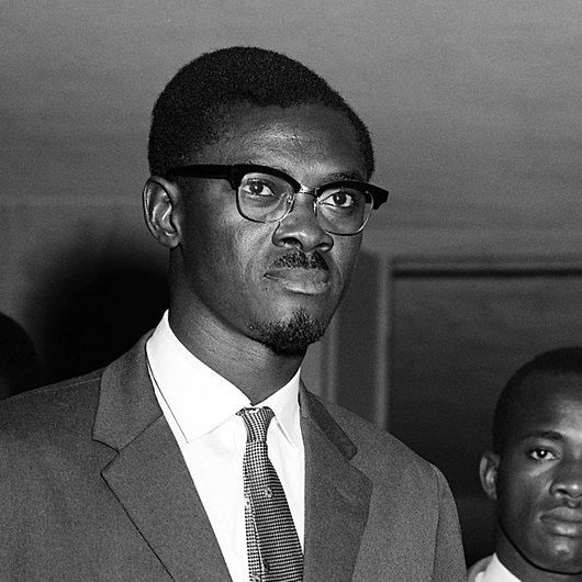 L'image montre un groupe d'hommes en costume, pris dans un cadre formel. Au premier plan, un jeune homme porte des lunettes à monture noire et affiche une expression sérieuse. Il est vêtu d'un costume sombre avec une chemise claire et une cravate. Derrière lui, deux autres hommes sont présents, également en costume, portant des expressions neutres. La lumière est douce, et l'atmosphère dégage un sentiment de gravité ou d'importance. Les murs autour d'eux semblent être peu décorés, mettant l'accent sur les personnes au centre de l'image.