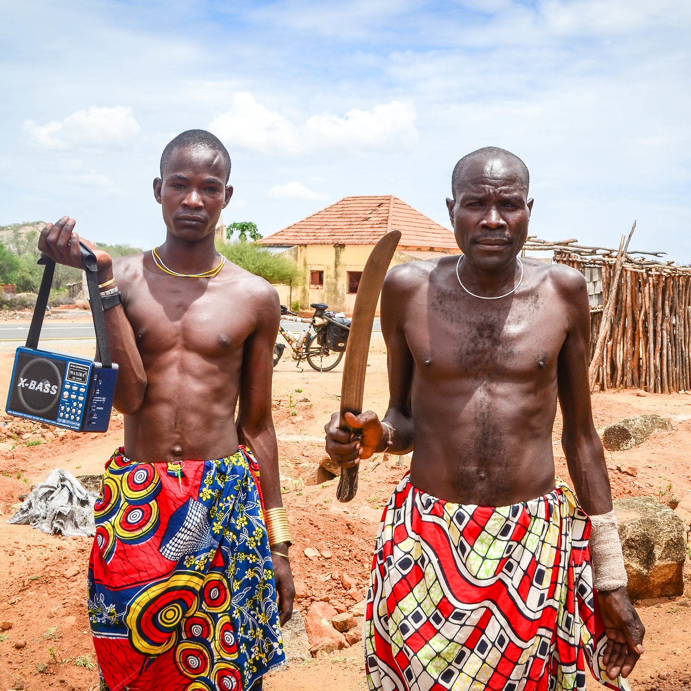 L'image montre deux hommes debout sur un terrain sec et rocheux. Ils portent des vêtements colorés avec des motifs vibrants, typiques d'une culture africaine. Le premier homme, à gauche, tient un poste de radio portable, tandis que le second, à droite, brandit une grande machette. En arrière-plan, on peut apercevoir des bâtiments simples, ainsi qu'une nature aride avec quelques arbres. Le ciel est bleu avec quelques nuages. L'atmosphère semble calme, mais la présence des objets et des vêtements indique une culture dynamique et vivante.
